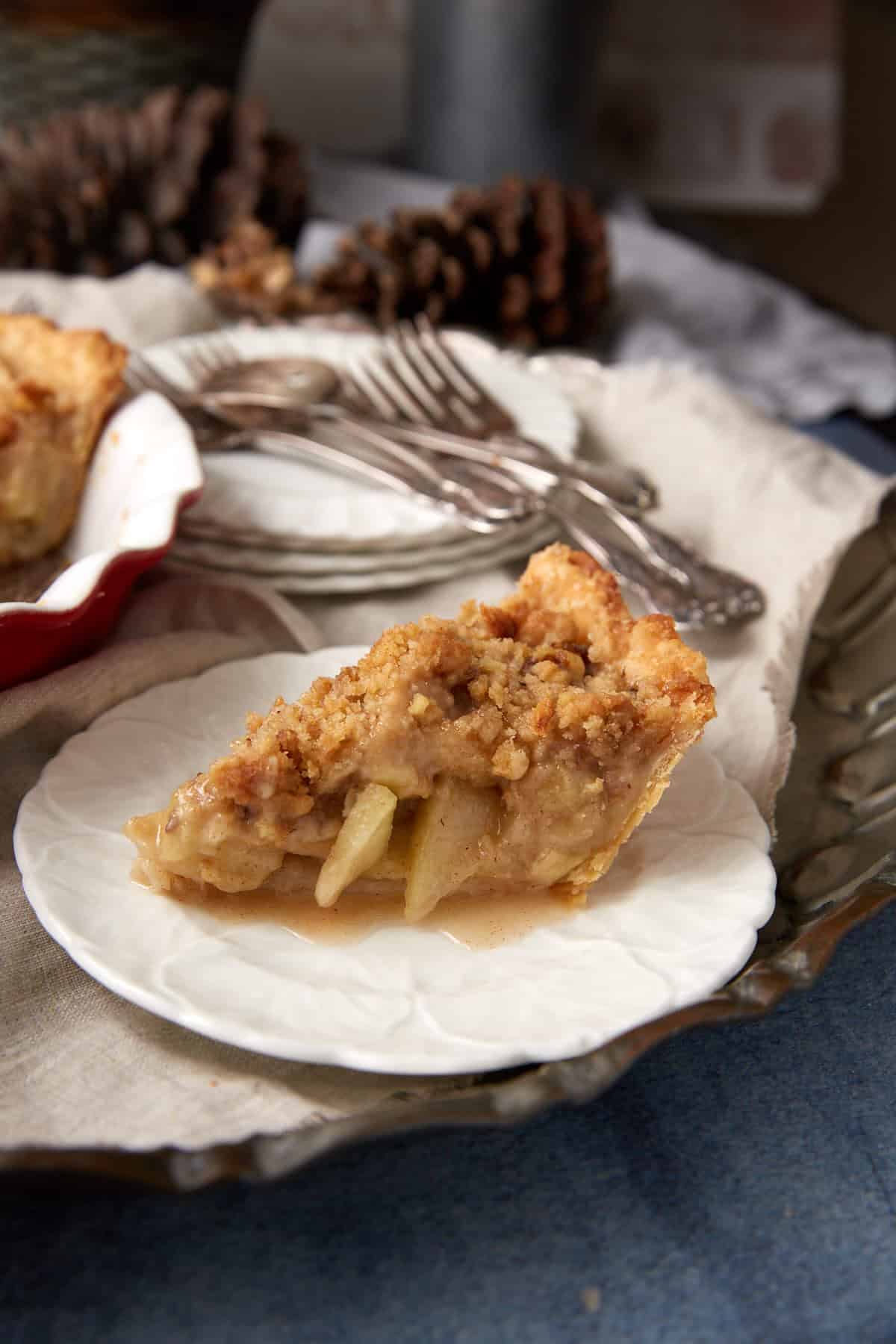 A slice of apple pie with crumb topping sits on a white plate, its filling and crumb layer visible. Behind it are forks, a pie dish with more pie, and pine cones for decoration—a tempting taste of the best Dutch apple pie recipe.