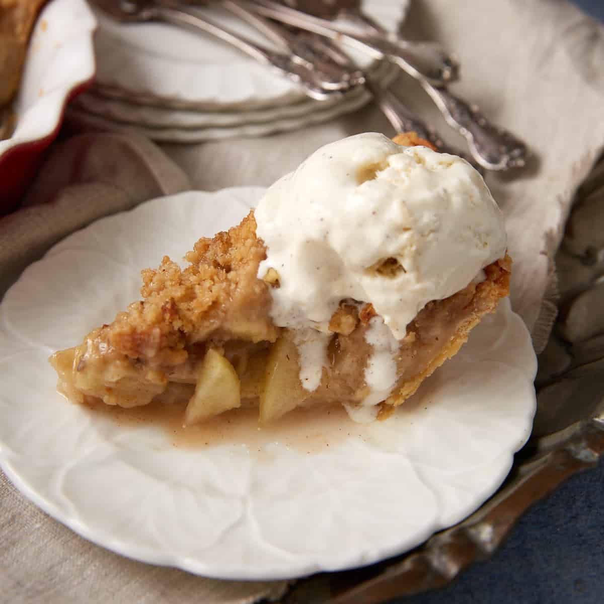 A slice of Apple Crumb Pie with a crumb topping is served on a white plate, topped with vanilla ice cream. Silverware rests on a napkin in the background, making this dessert look like the best apple crumb pie recipe come to life.