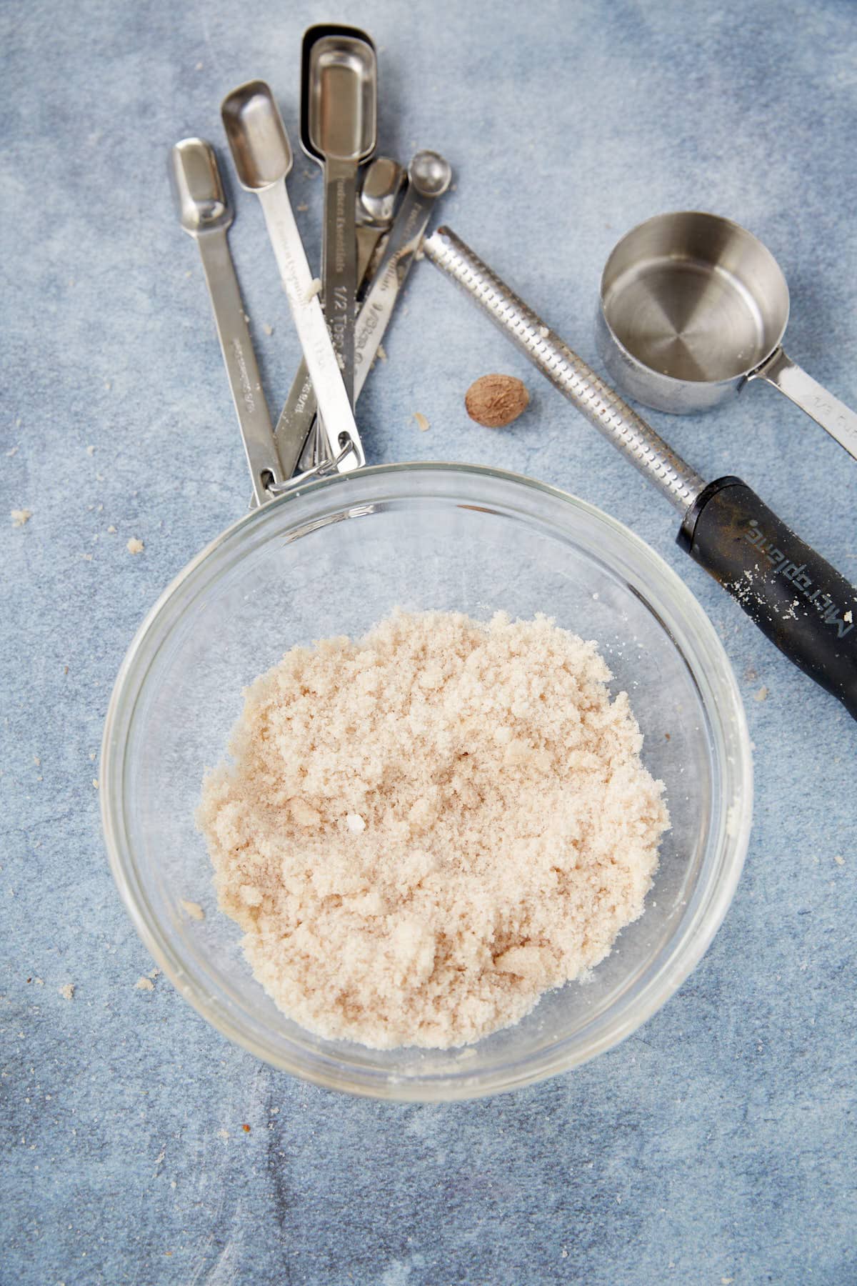 A glass bowl filled with a crumbly, beige mixture—perfect for an apple pie with crumb topping—sits on a blue surface. Surrounding the bowl are measuring spoons, a metal measuring cup, a whole nutmeg, and a microplane grater.