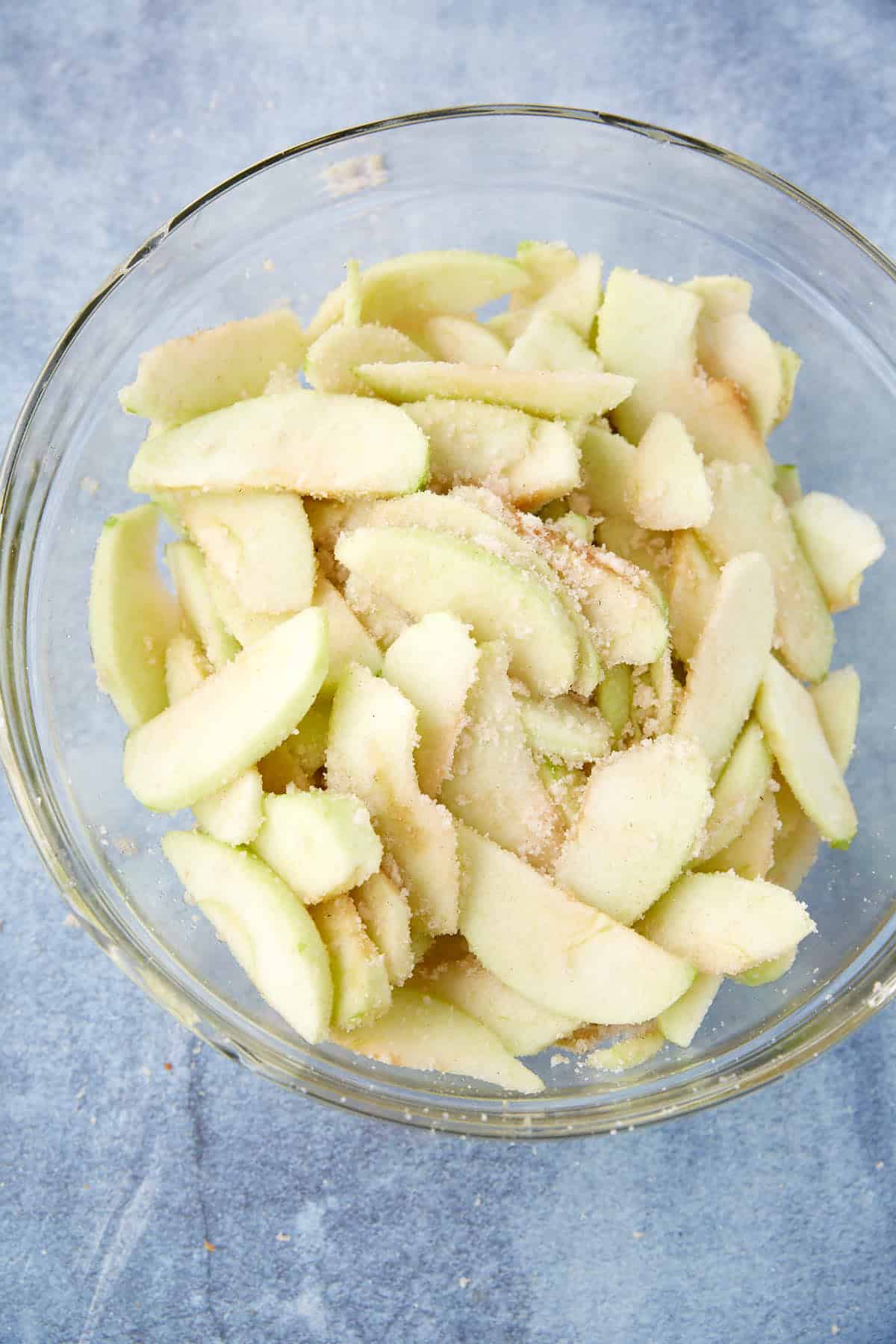 A glass bowl filled with thin slices of peeled green apples coated in sugar and spices, ready to be used for a delicious apple crumble pie, sits on a light blue surface.