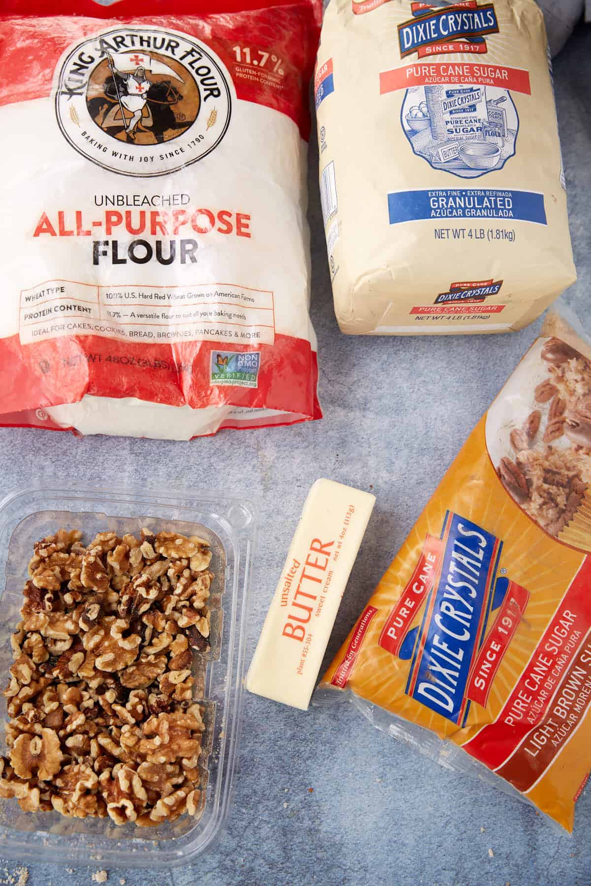 Bags of flour and sugar, a stick of butter, and chopped walnuts are ready on a blue-gray surface—essential ingredients for making the best apple crumb pie recipe or a classic apple pie with crumb topping.