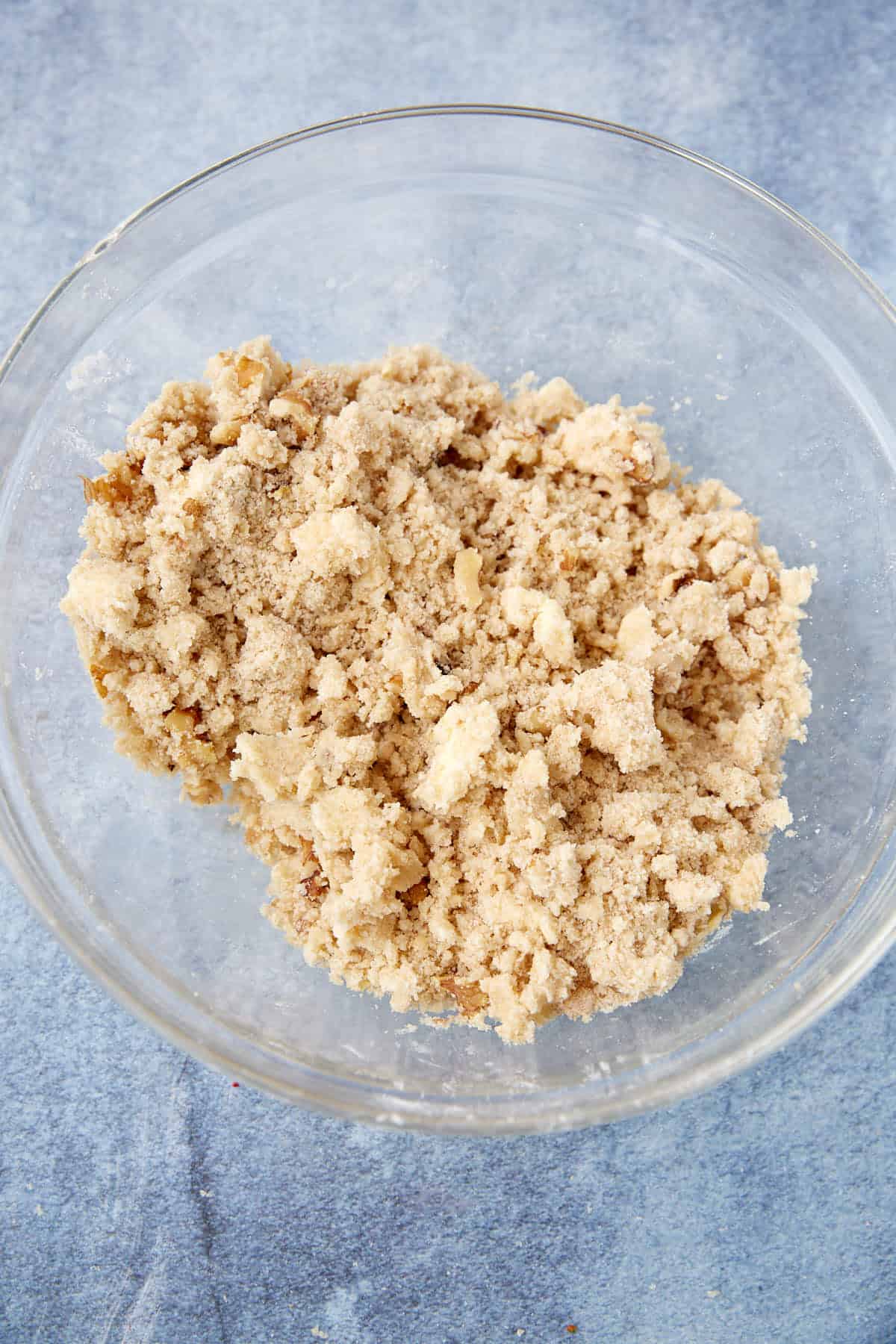 A clear glass bowl containing a crumbly mixture of flour, sugar, and butter sits on a light blue surface—perfect for making an Apple Crumb Pie or adding to your favorite apple pie with crumb topping.