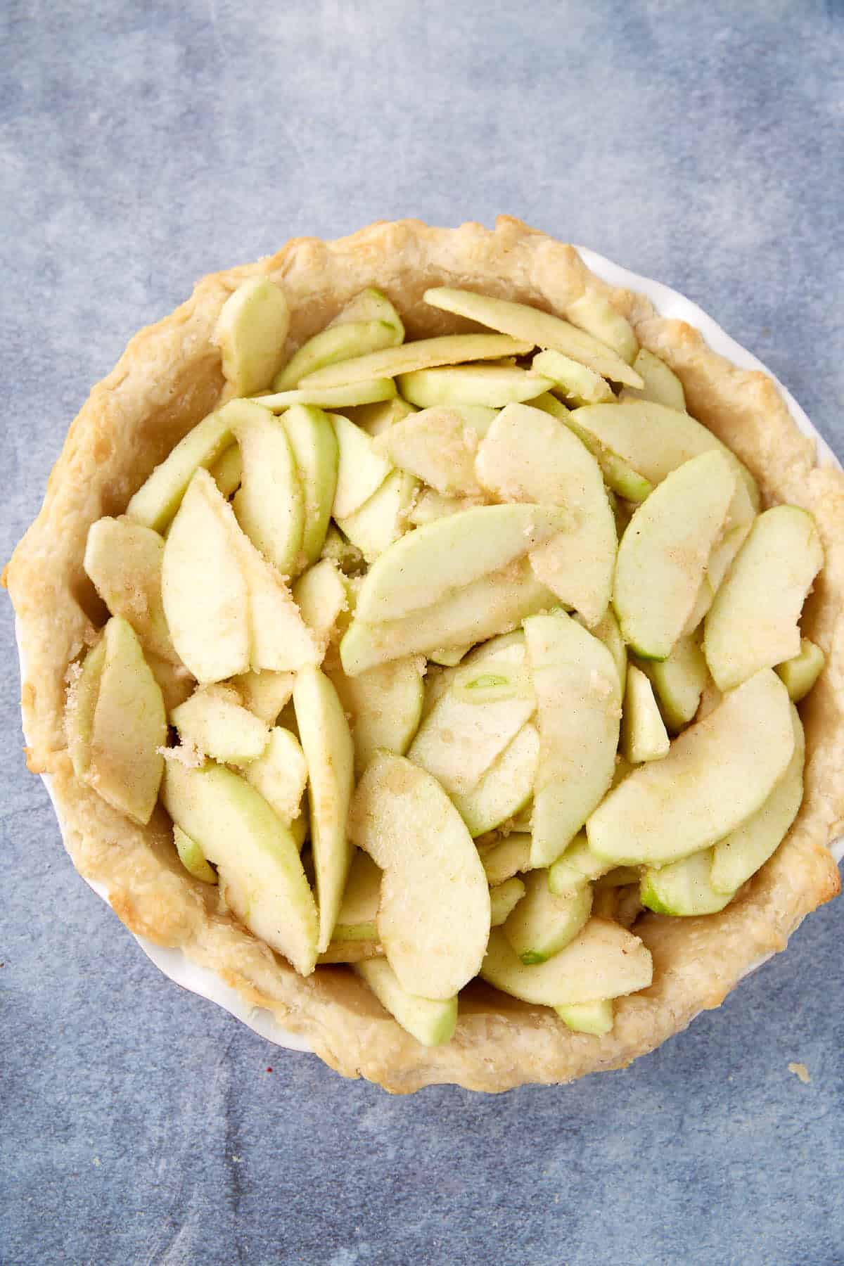 A pie crust filled with raw, sliced green apples coated in sugar, ready to be baked into the best apple crumb pie recipe, sits on a gray textured surface.