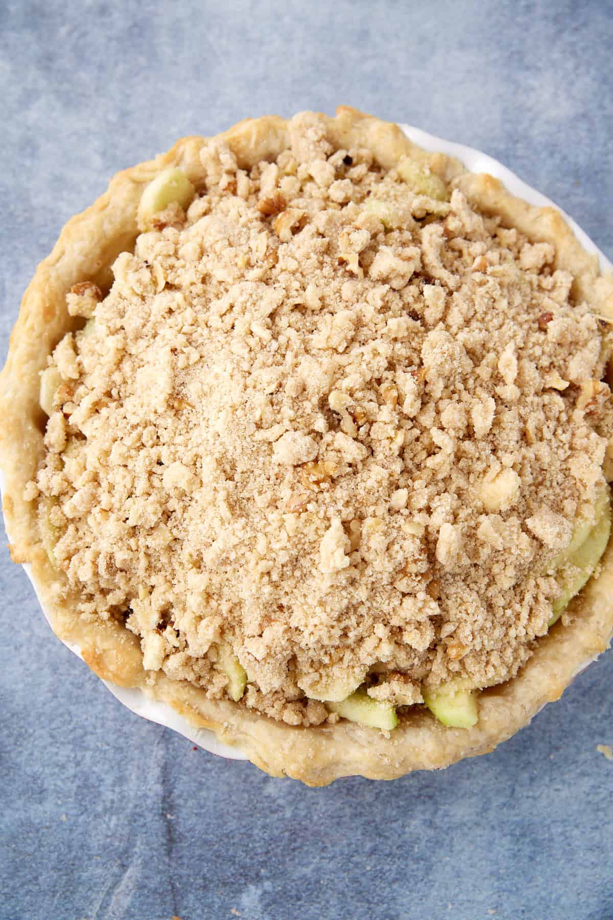 A baked Apple Crumb Pie topped with a generous layer of golden-brown crumbly streusel sits on a blue textured surface. The pie crust and streusel topping appear crispy and homemade.