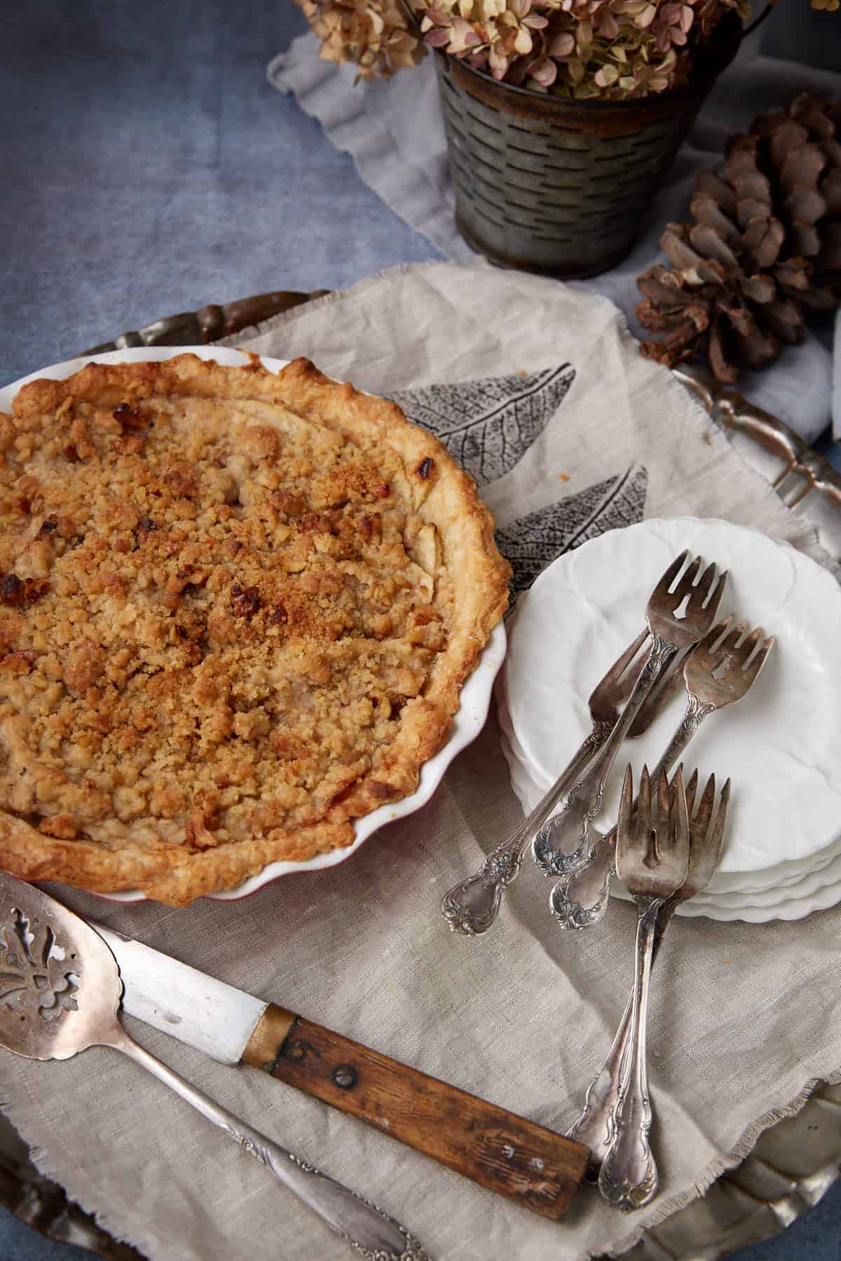 An apple crumble pie in a white dish sits on a tray with a pie server, dessert forks on small plates, and a linen napkin. Dried flowers and pinecones decorate the background, evoking a rustic autumn setting.