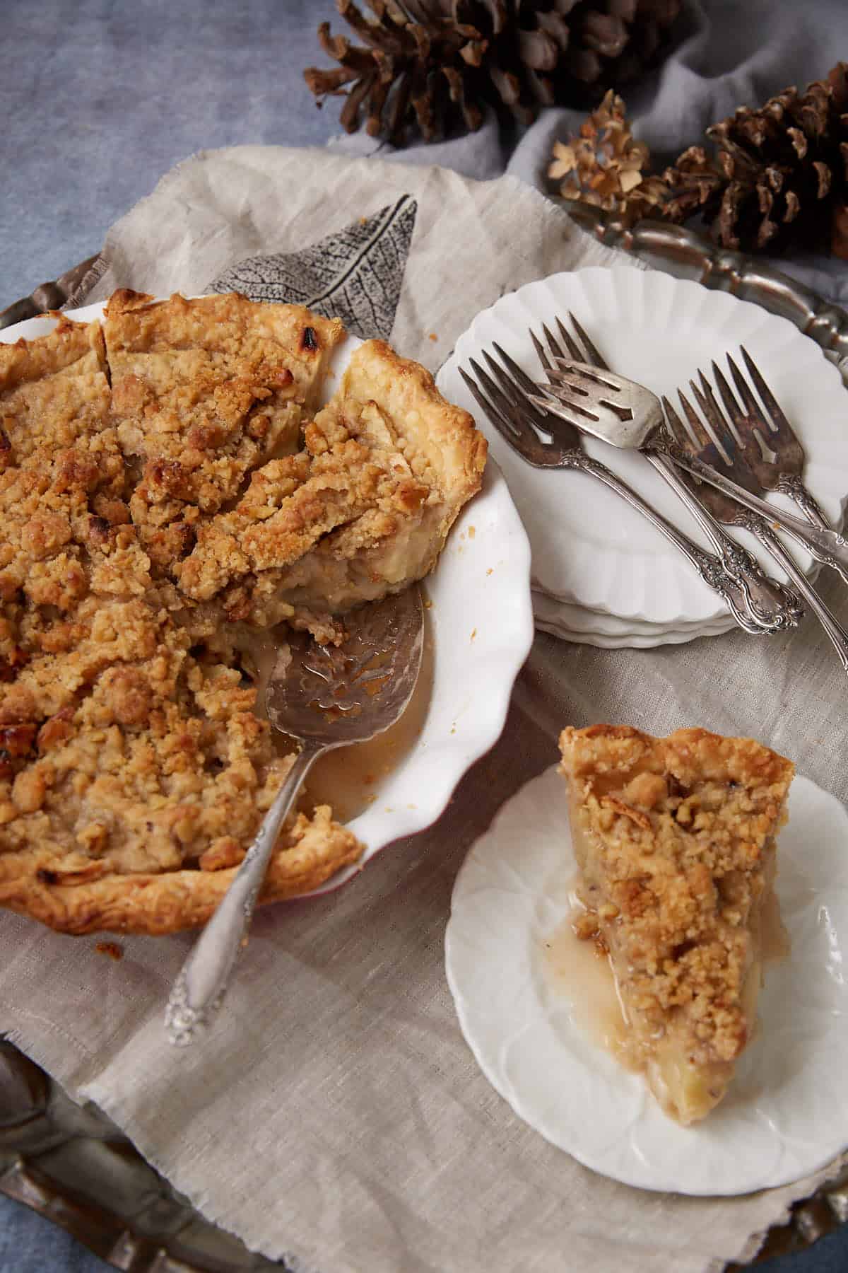 A slice of apple pie with crumb topping rests on a small plate beside a partially served pie in a white dish. Silver forks and pine cones are arranged nearby on a linen cloth, highlighting the cozy appeal of Apple Crumb Pie.