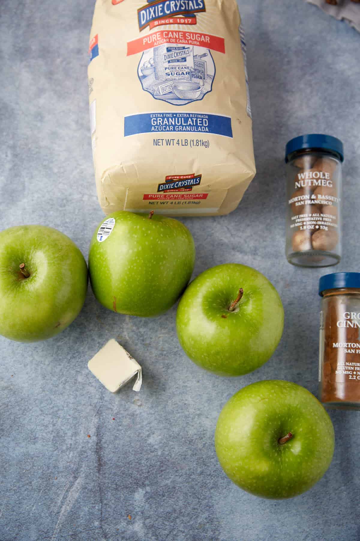 Three green apples, a stick of butter, granulated sugar, whole nutmeg, and ground cinnamon are arranged on a gray surface—classic ingredients for the best apple crumb pie recipe.