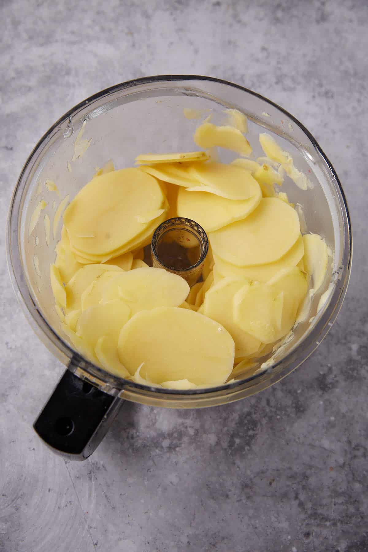 A food processor bowl filled with thinly sliced raw potatoes, ready to be transformed into creamy scalloped potatoes, sits on a gray, textured surface.