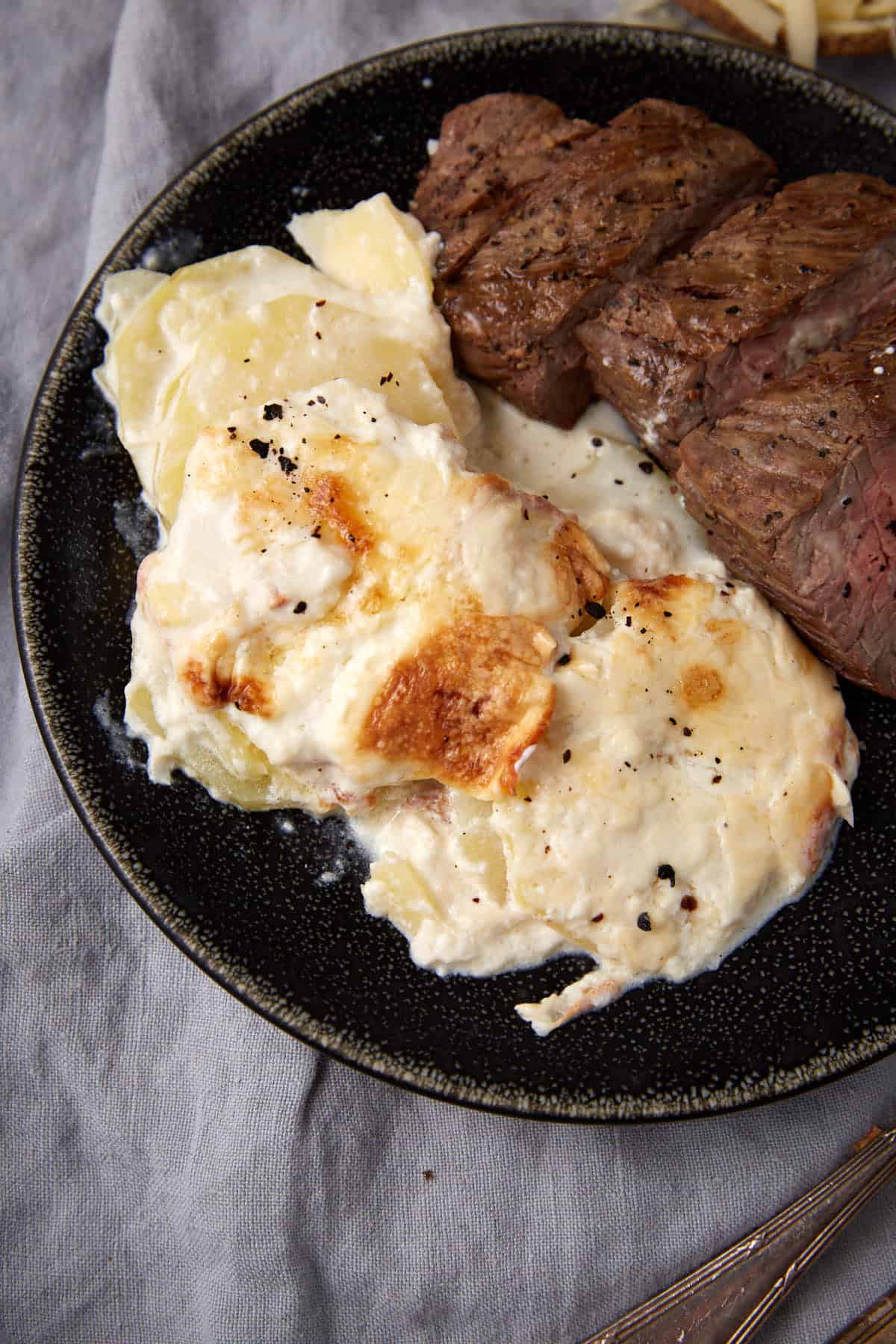 A plate with creamy scalloped potatoes with cheese, topped with browned cheese, sits beside slices of grilled steak on a dark plate with a gray cloth underneath.