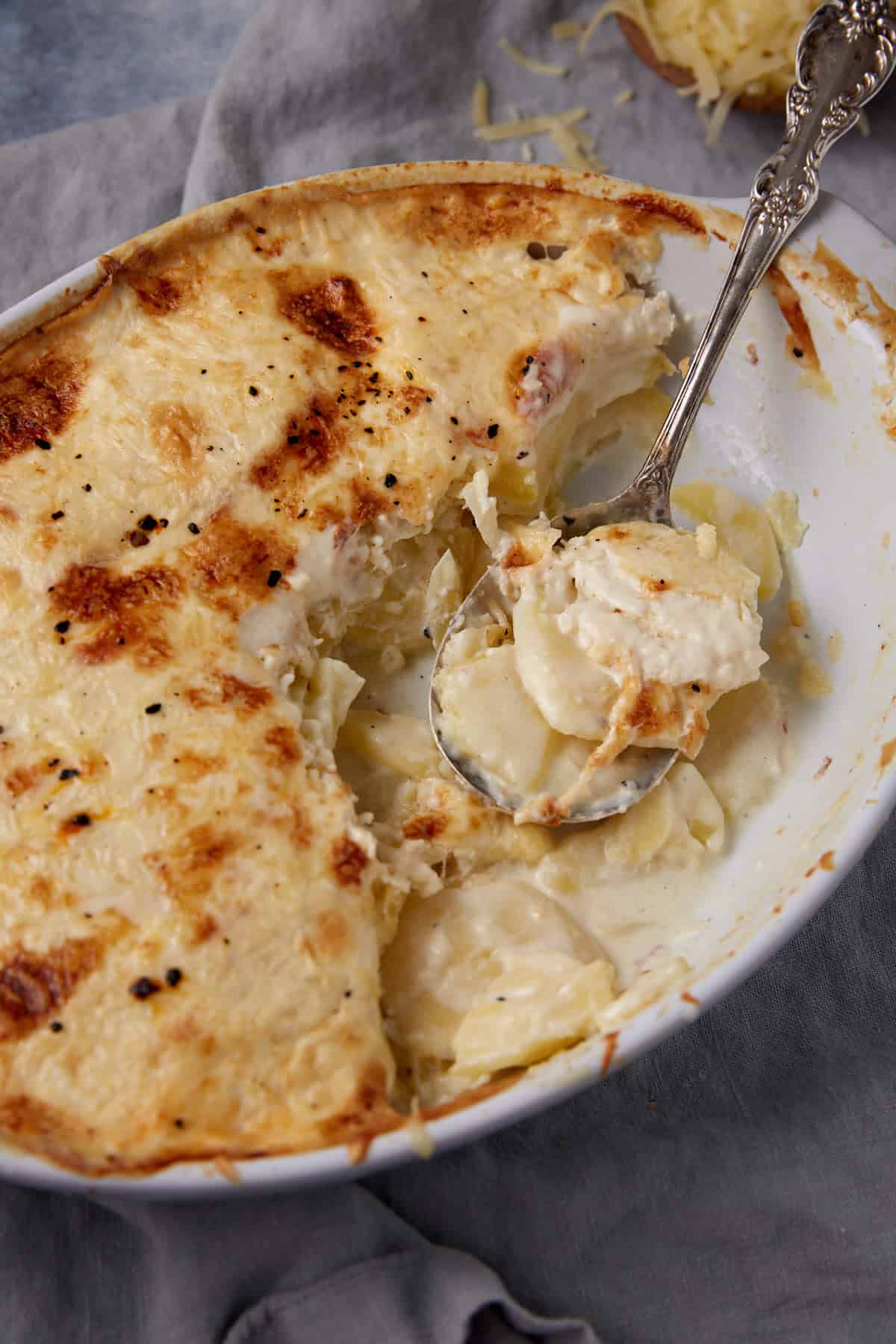 A baked dish of creamy scalloped potatoes with a golden brown cheesy top. A serving spoon rests in the dish, holding a portion of thinly sliced potatoes in a rich sauce. Some cheese and sauce are spilled on the edge.