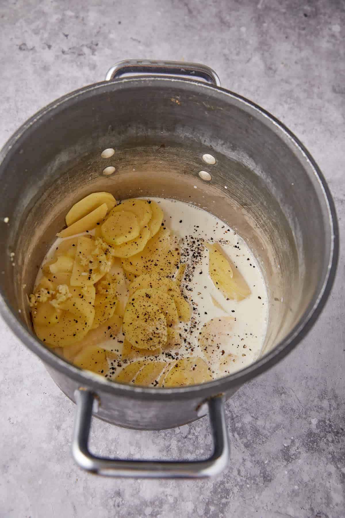 Sliced potatoes, minced garlic, milk, and ground black pepper in a metal pot on a gray countertop—everything you need to start delicious homemade scalloped potatoes, ready to be cooked.