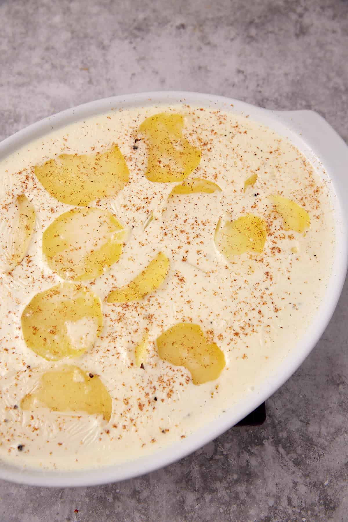 An oval white dish filled with creamy homemade scalloped potatoes, sprinkled with ground black pepper and nutmeg, ready to be baked, sitting on a gray textured surface.
