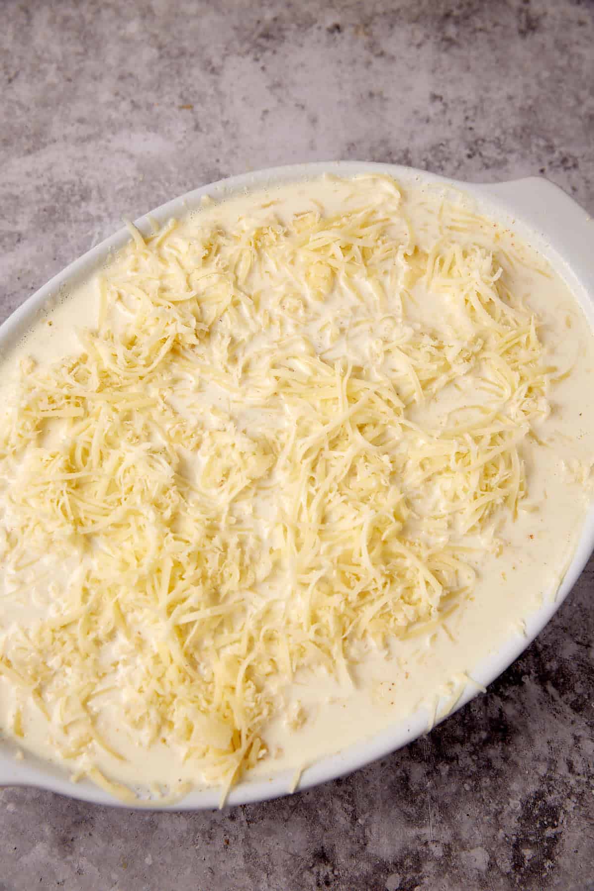A white oval baking dish filled with creamy scalloped potatoes, topped with shredded cheese and rich sauce, sits ready to be baked on a textured gray countertop.