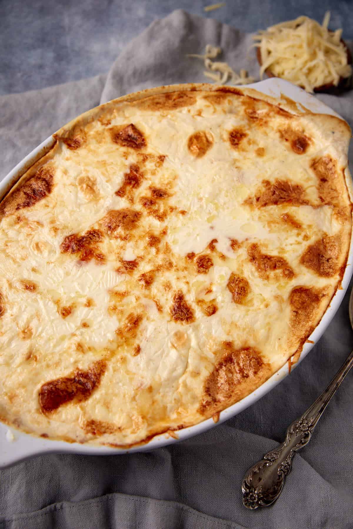 A baked casserole dish filled with golden-brown, cheesy homemade scalloped potatoes with cream sits on a gray cloth, with shredded cheese and an ornate silver serving spoon nearby.