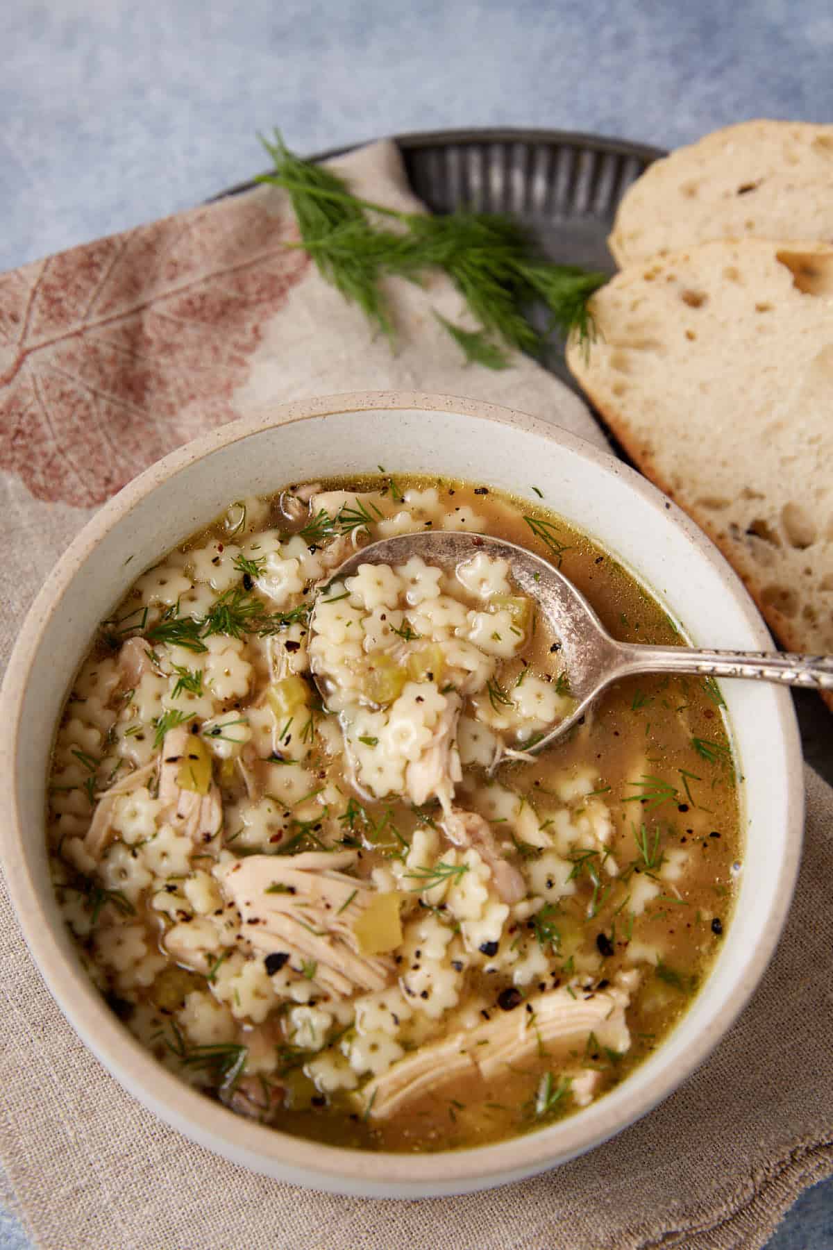 A bowl of Chicken Orzo Soup with lemon, featuring star-shaped pasta, shredded chicken, fresh herbs, and broth sits on a napkin beside slices of rustic bread. A spoon rests in the bowl, ready for eating.