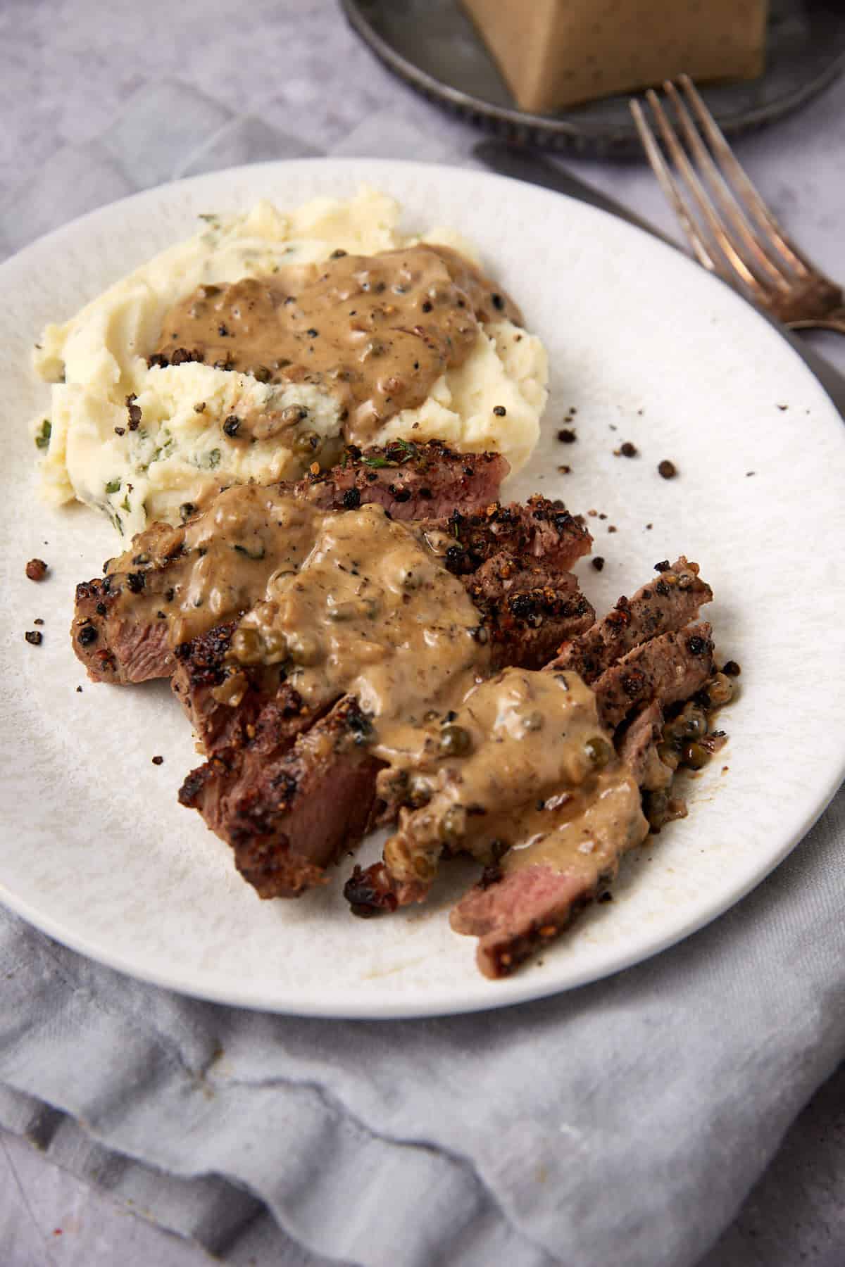 Sliced steak au poivre topped with peppercorn sauce served next to creamy mashed potatoes on a white plate, with a fork and a piece of bread visible in the background.