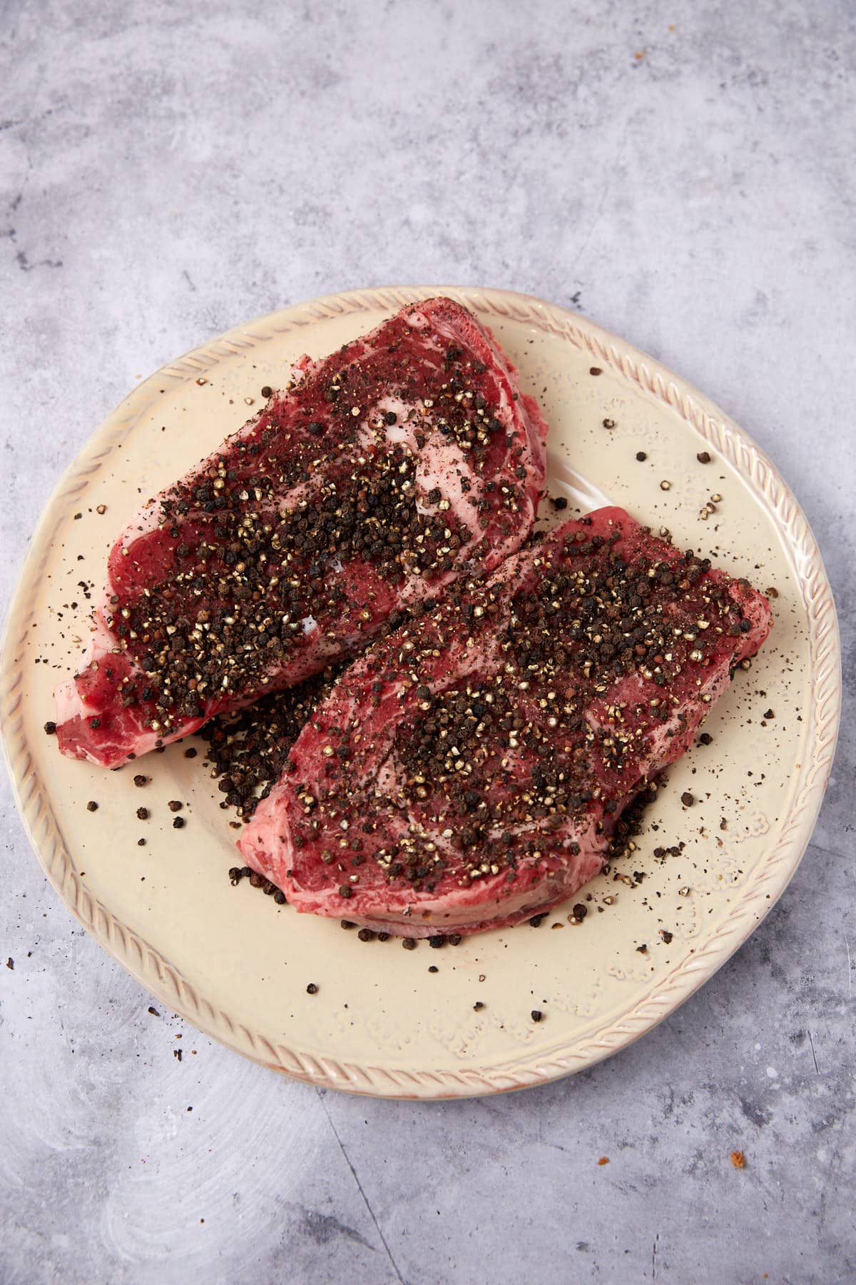 Two raw steaks on a cream-colored plate, generously coated with coarse black pepper and seasonings, set on a light textured surface—perfect for preparing an authentic au poivre sauce recipe.