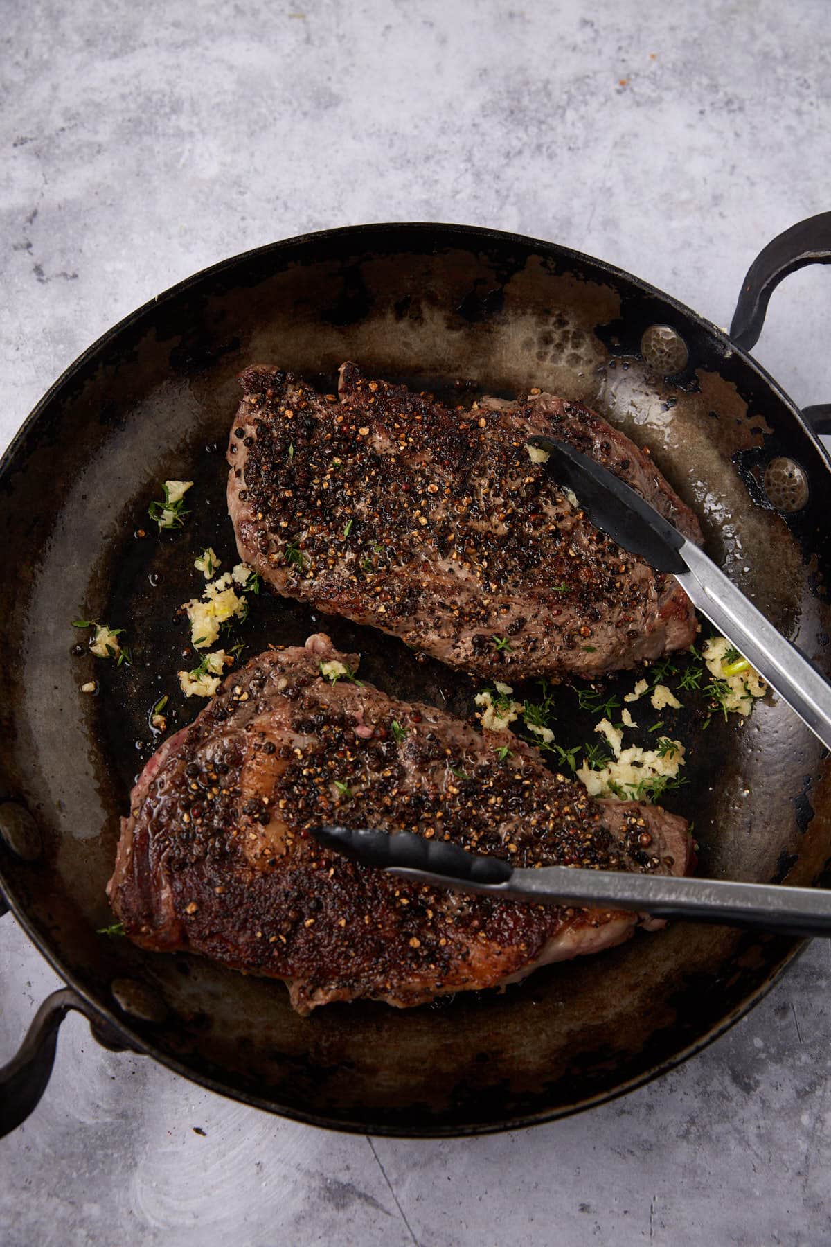 A cast iron skillet with two seasoned, cooked steaks topped with minced garlic and herbs. A pair of metal tongs rests on one steak, ready to serve alongside a rich creamy steak sauce, on a light gray textured surface.