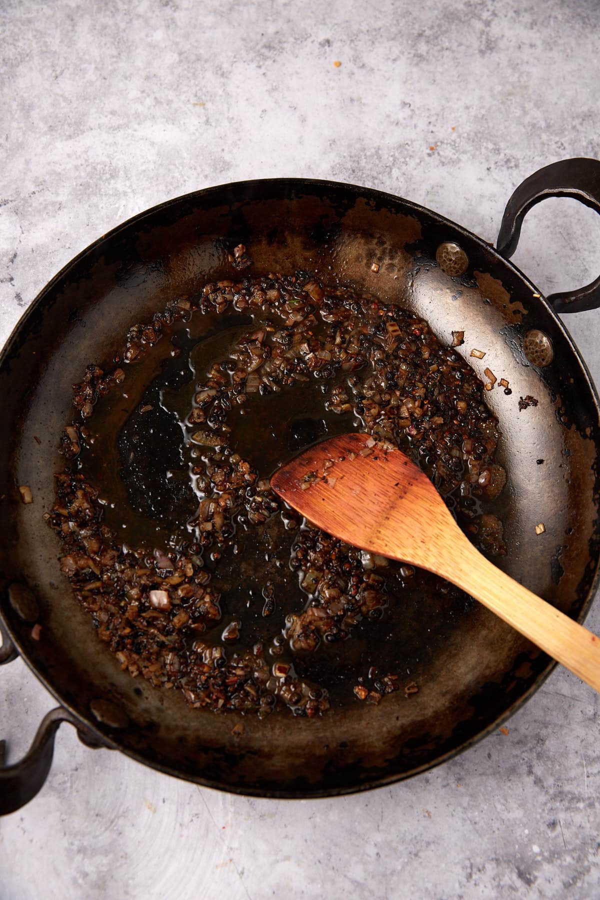 A black metal pan with sautéed, browned onions and oil residue sits on a gray surface. A wooden spatula rests inside, suggesting the start of a rich au poivre sauce recipe for a creamy steak sauce. The pan has dark handles on both sides.