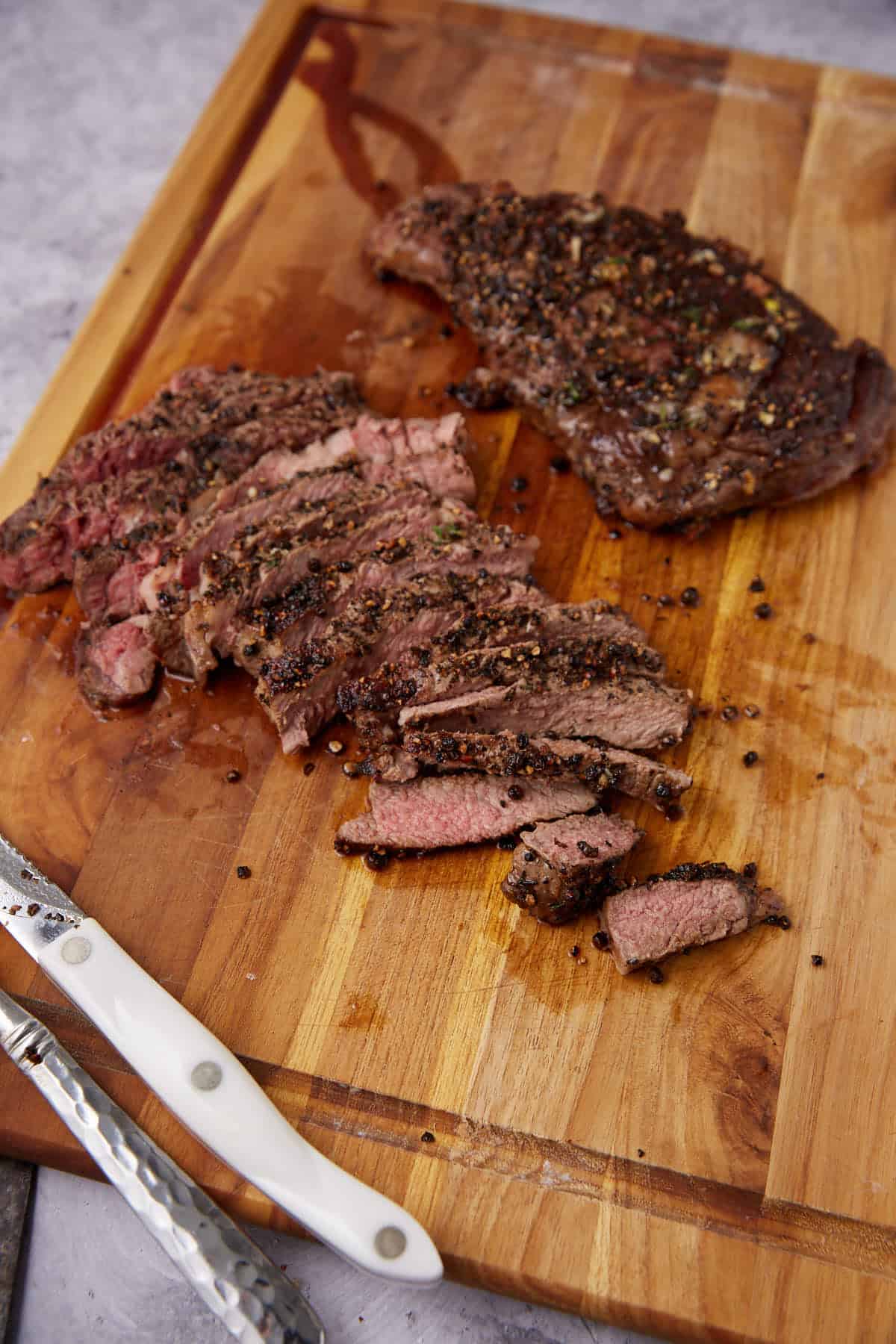 A cooked steak, partly sliced, sits on a wooden cutting board. The meat is seasoned with cracked pepper and herbs in classic au poivre style. A steak knife and fork with white handles rest nearby.