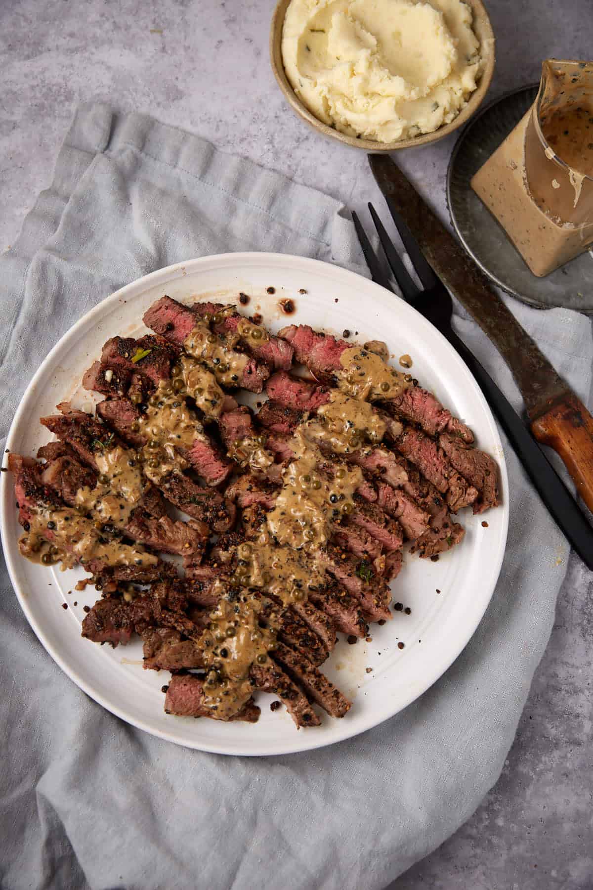 Sliced steak au poivre topped with peppercorn sauce on a white plate, accompanied by a fork and knife. In the background, there&rsquo;s a small bowl of mashed potatoes and a block of sauce, all set on a gray cloth.