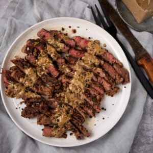 Sliced steak cooked medium-rare, arranged on a white plate and topped with a classic steak au poivre creamy sauce. A fork and knife rest nearby on a light gray cloth.