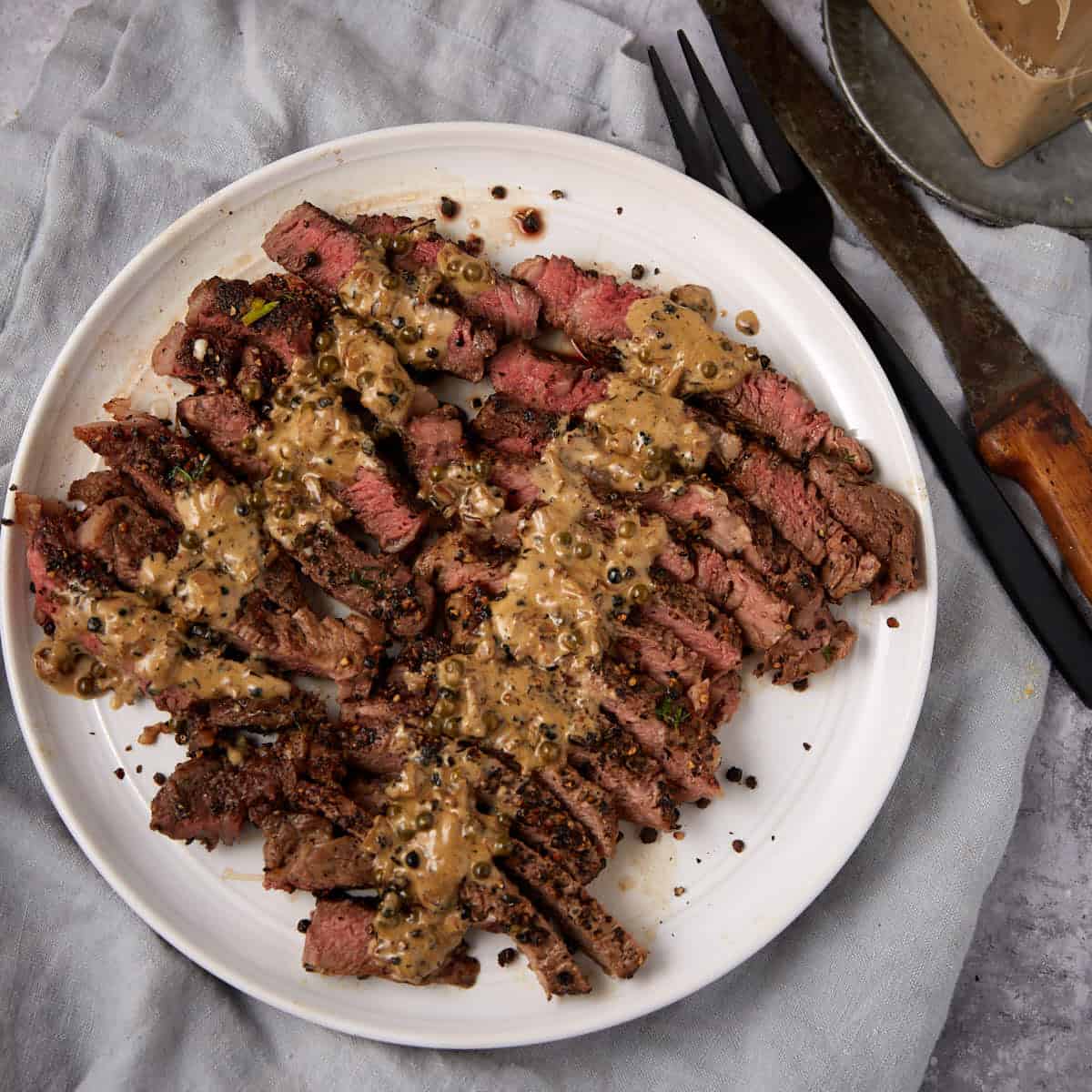 Sliced steak cooked medium-rare, arranged on a white plate and topped with a classic steak au poivre creamy sauce. A fork and knife rest nearby on a light gray cloth.