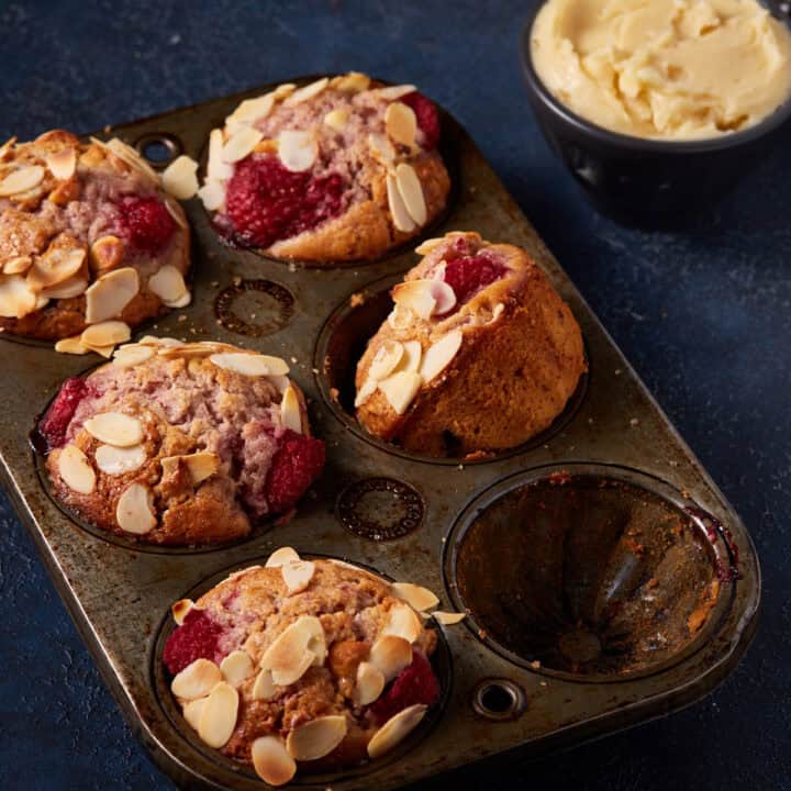 Raspberry white chocolate muffins in a muffin tin next to a bowl of honey butter.
