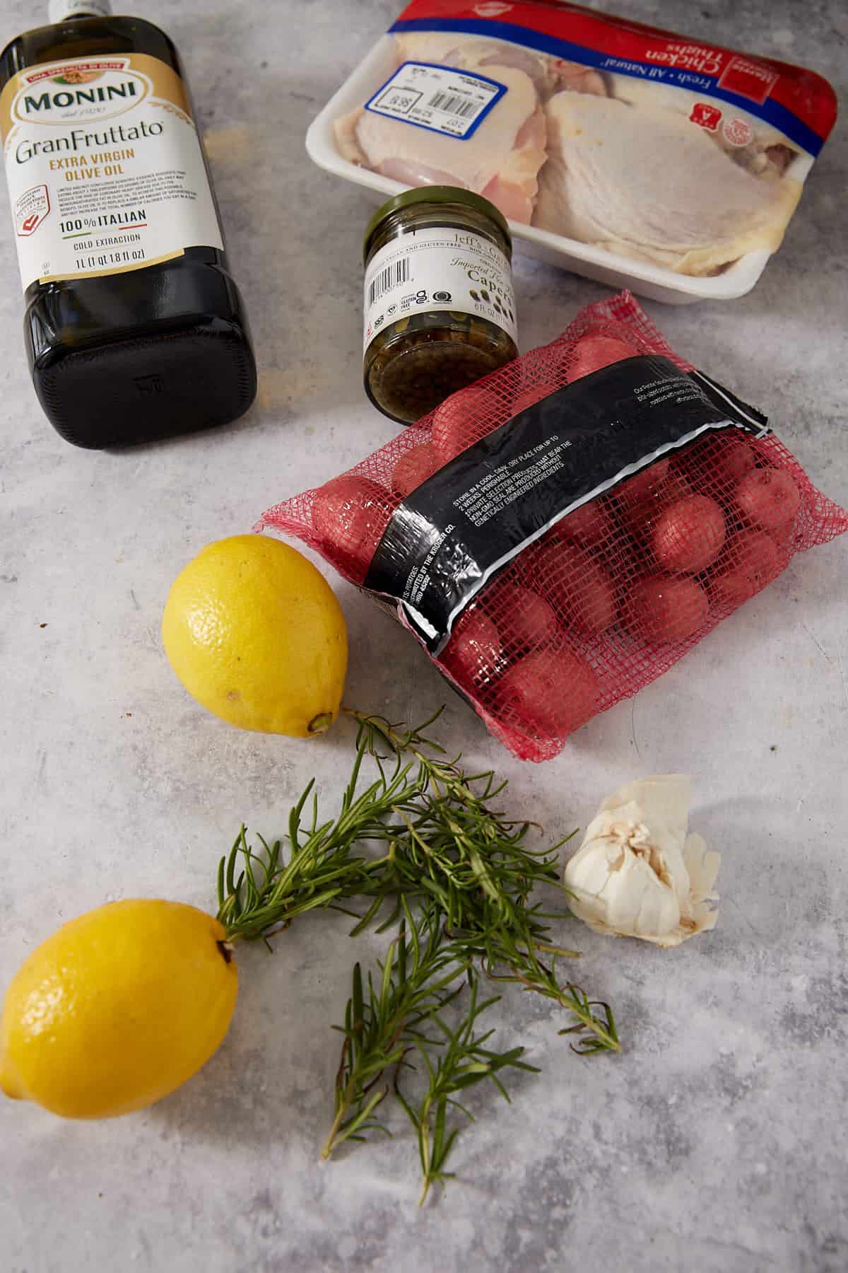 A bottle of olive oil, jar of pesto, pack of chicken, bag of red potatoes, two lemons, a sprig of rosemary, and a bulb of garlic rest on a gray surface—everything you need for an easy lemon chicken recipe or classic lemon chicken and potatoes.