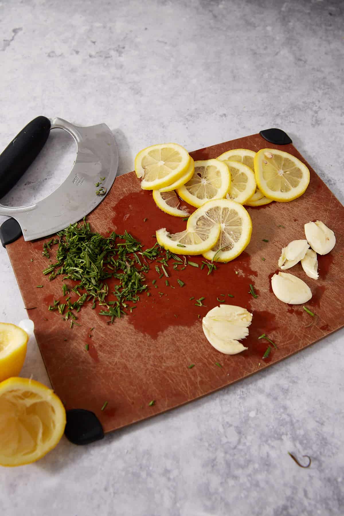 A cutting board with sliced lemon, chopped herbs, garlic cloves, and a mezzaluna knife on a gray countertop—perfect for prepping ingredients for baked lemon chicken or your favorite lemon chicken thighs recipe.