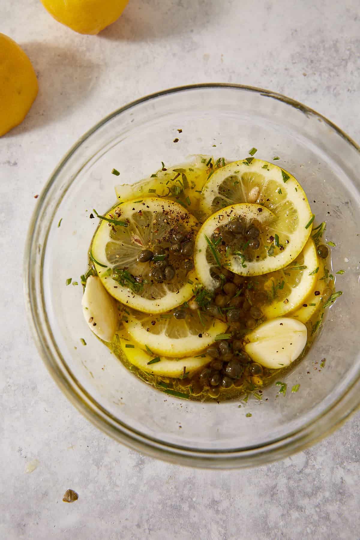 A glass bowl filled with sliced lemons, olive oil, capers, garlic cloves, chopped herbs, and black pepper—perfect ingredients for preparing flavorful lemon chicken thighs. Whole lemons sit nearby on a light grey countertop.