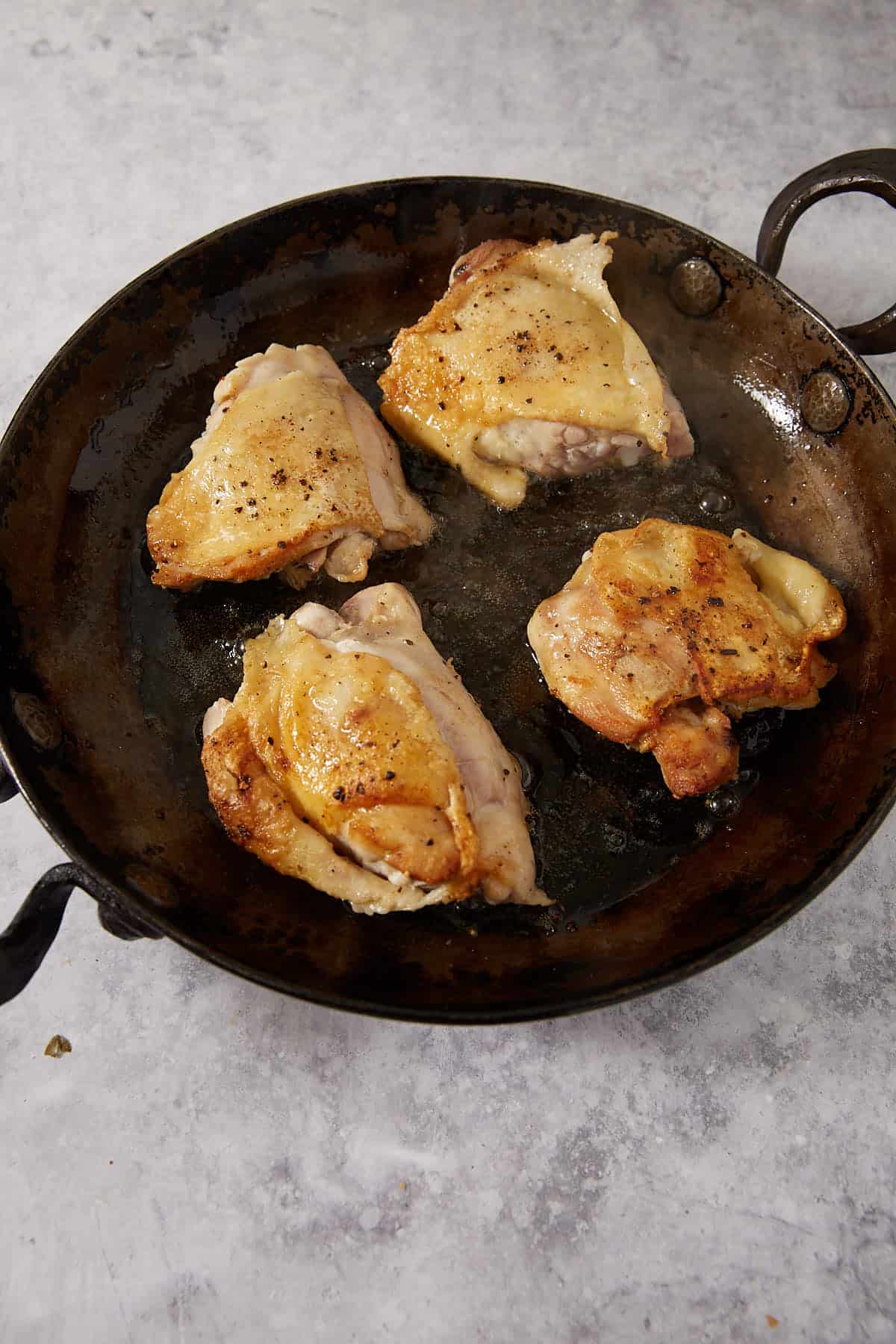 Four pieces of chicken with golden, crispy skin are cooking in a dark, round skillet. The pan sits on a light gray, textured surface. The chicken is seasoned with salt and pepper, embodying the essence of an easy lemon chicken recipe. Steam rises from the hot pan, promising mouthwatering lemon baked chicken ready to be enjoyed.