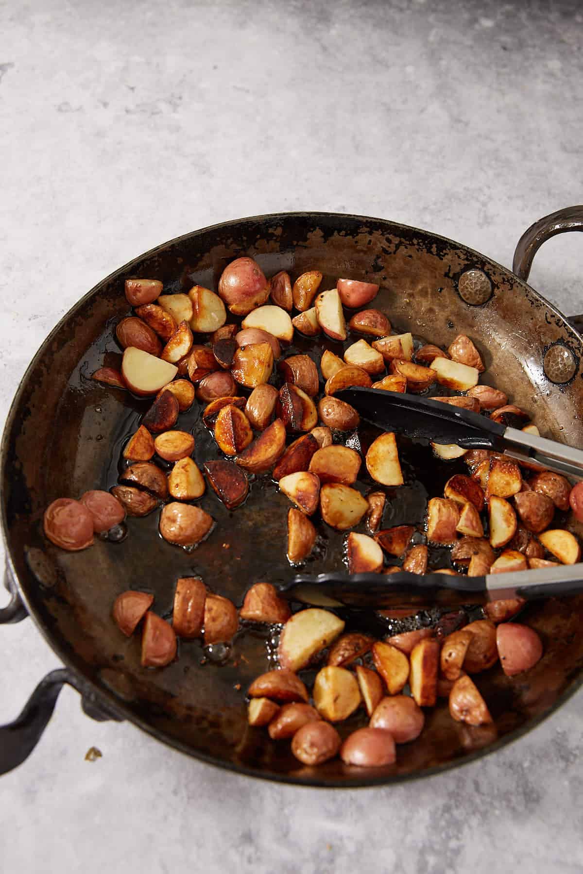 Sautéed, sliced baby potatoes are being cooked in a large, rustic skillet alongside juicy lemon chicken thighs. Metal tongs rest on the edge, ready to stir. The potatoes are golden brown and crispy, a perfect side for this easy lemon chicken recipe.