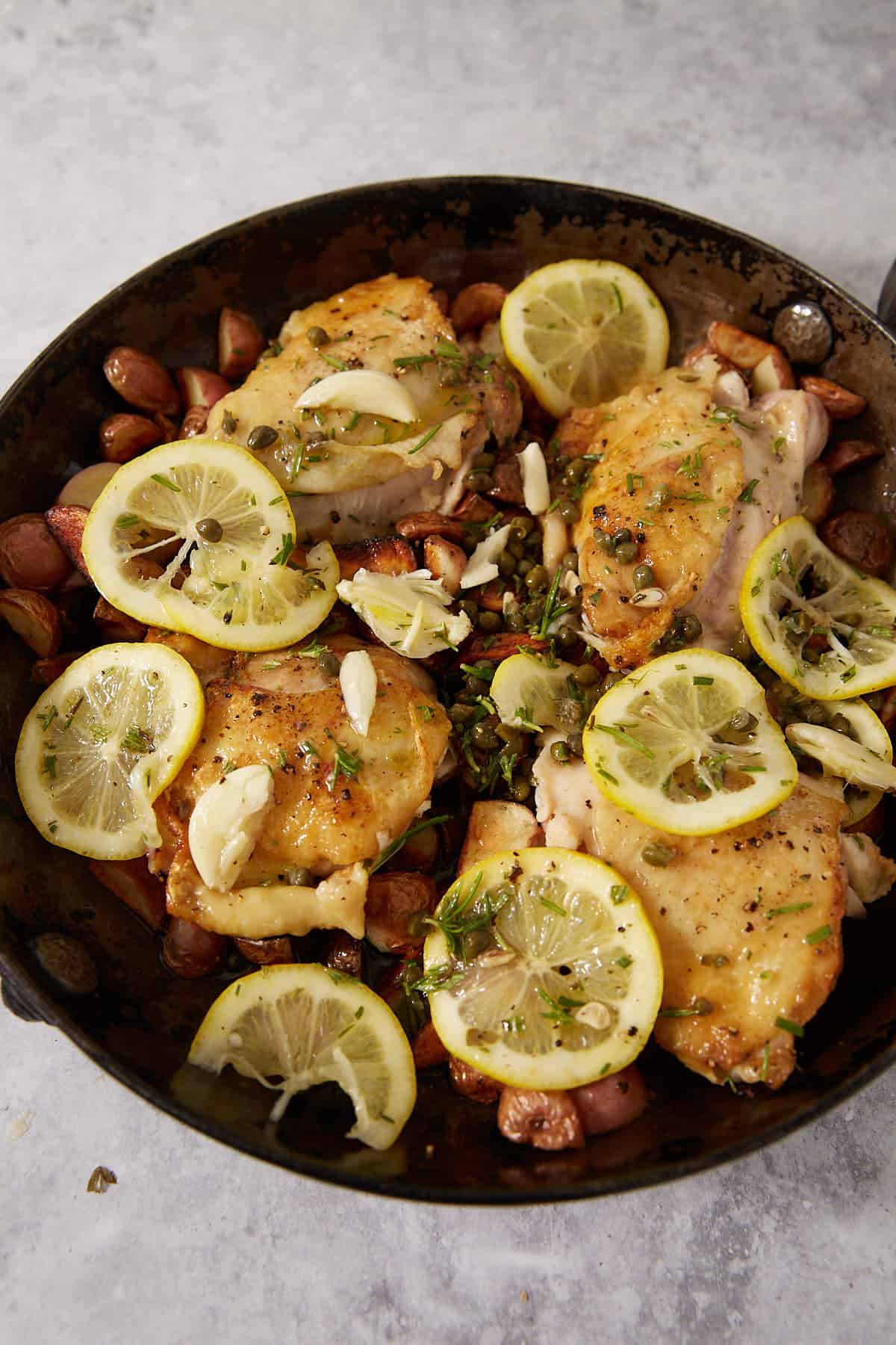 Chicken thighs cooked in a skillet with sliced lemons, garlic cloves, herbs, grapes, and capers on a light gray surface—this easy lemon chicken recipe brings bright citrus flavor and savory notes together for a delicious meal.
