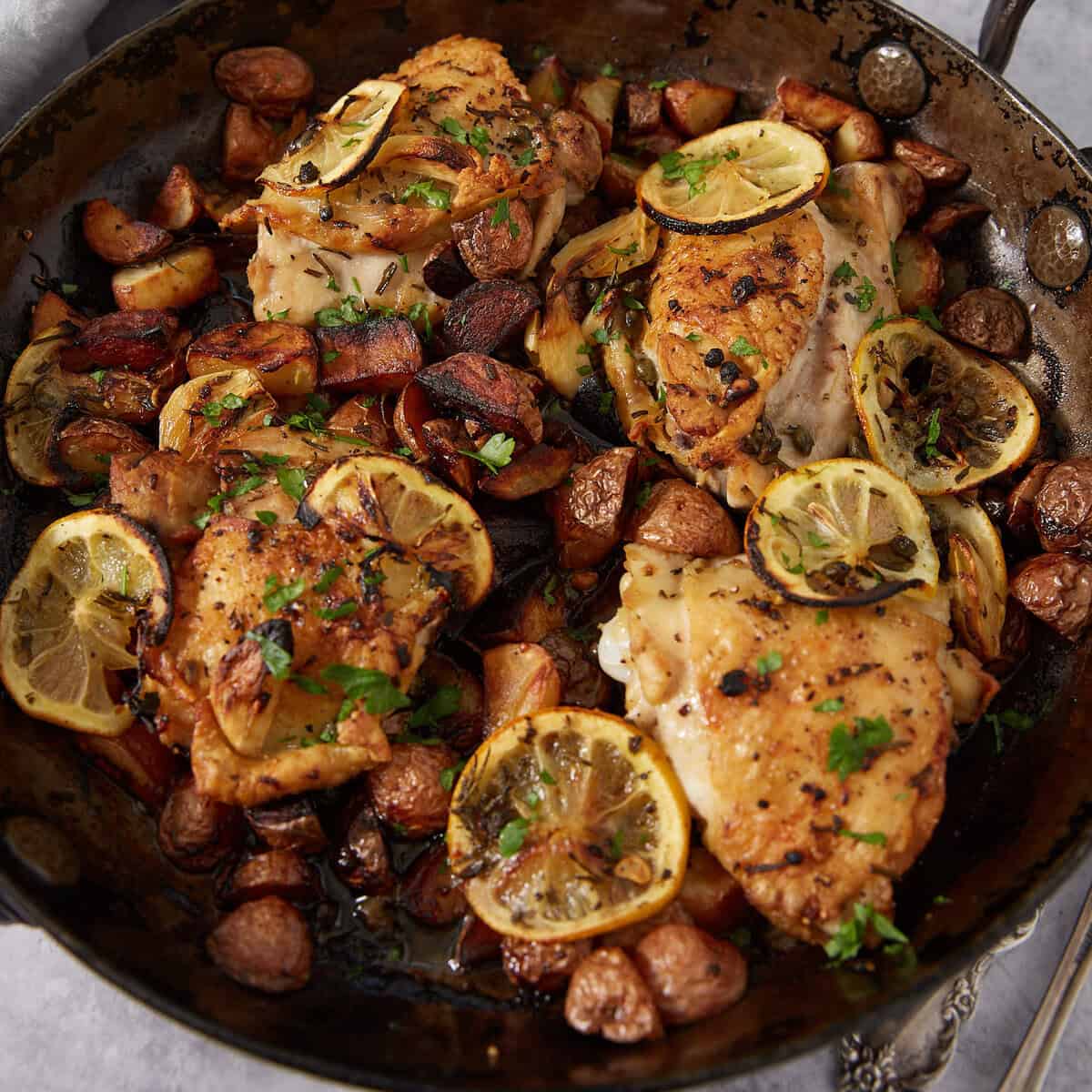 Baked lemon chicken thighs with sliced lemons and golden potatoes, garnished with chopped herbs and served in a rustic skillet—an easy lemon chicken recipe perfect for any cozy meal.