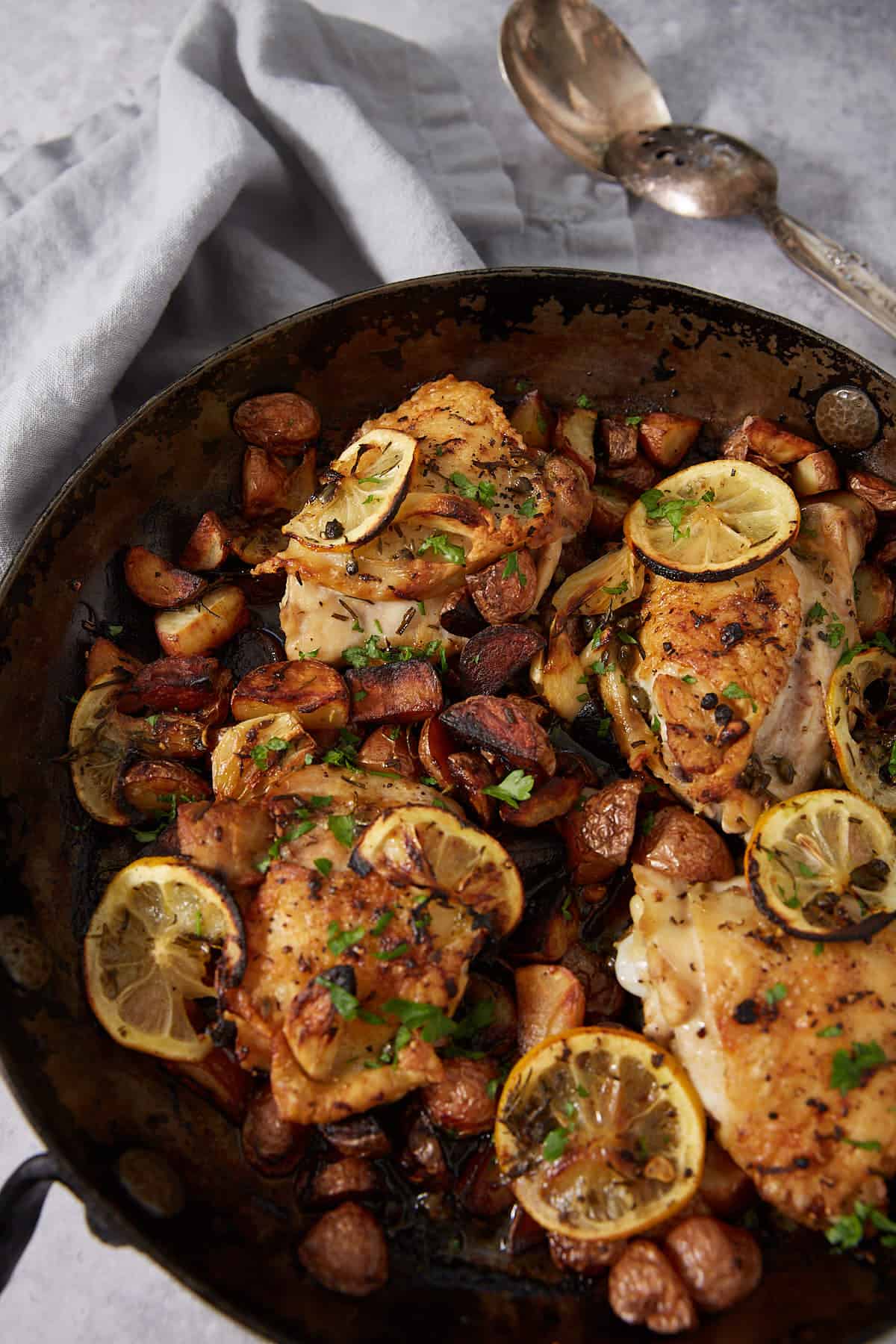 A skillet filled with baked lemon chicken thighs, sliced lemons, and diced potatoes, garnished with herbs. A gray napkin and serving utensils are visible in the background.