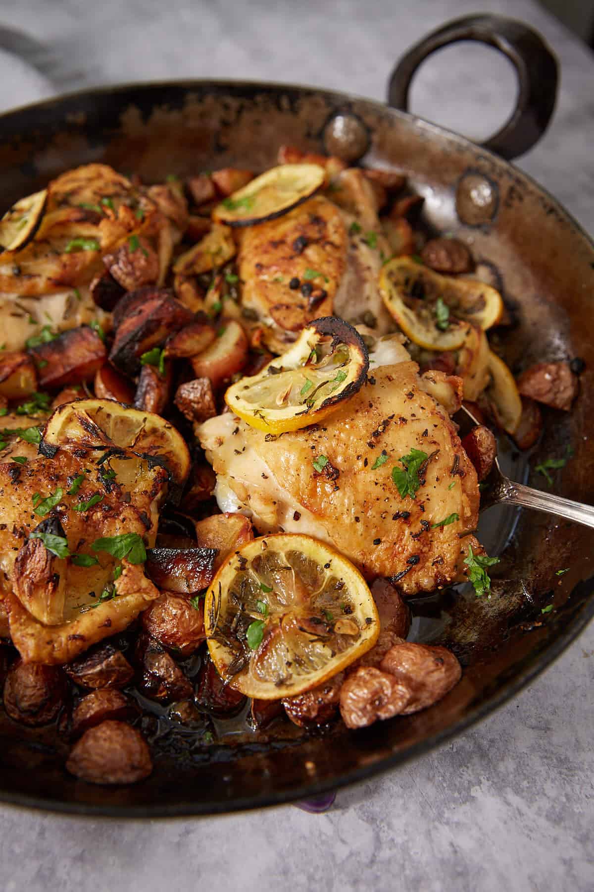 A rustic pan filled with juicy lemon chicken thighs topped with fresh lemon slices, surrounded by roasted potatoes and herbs on a gray surface—an easy lemon chicken recipe perfect for weeknight dinners.