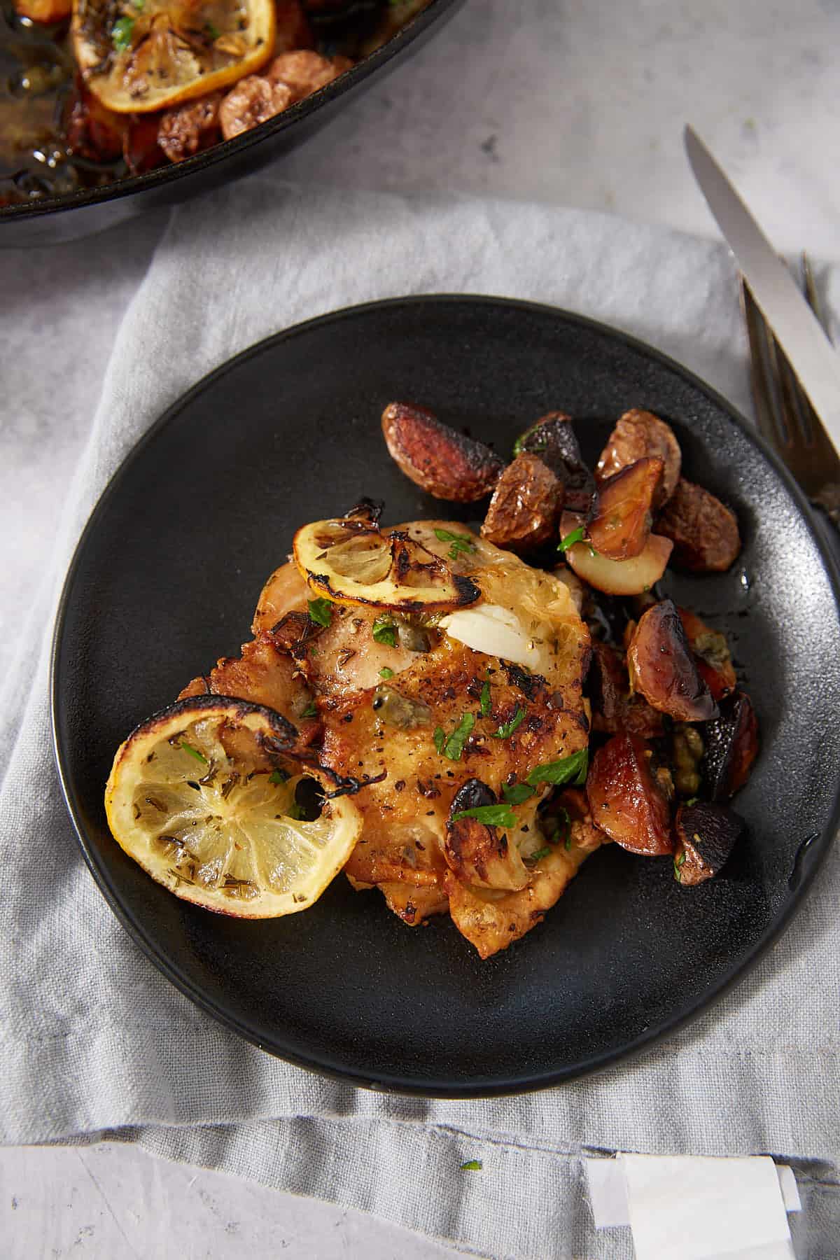 A black plate with roasted chicken garnished with lemon slices, herbs, and garlic, served alongside roasted figs and vegetables on a gray cloth napkin. Perfect for those seeking an easy lemon chicken recipe, this dish is elegantly presented with a fork and knife placed to the right.