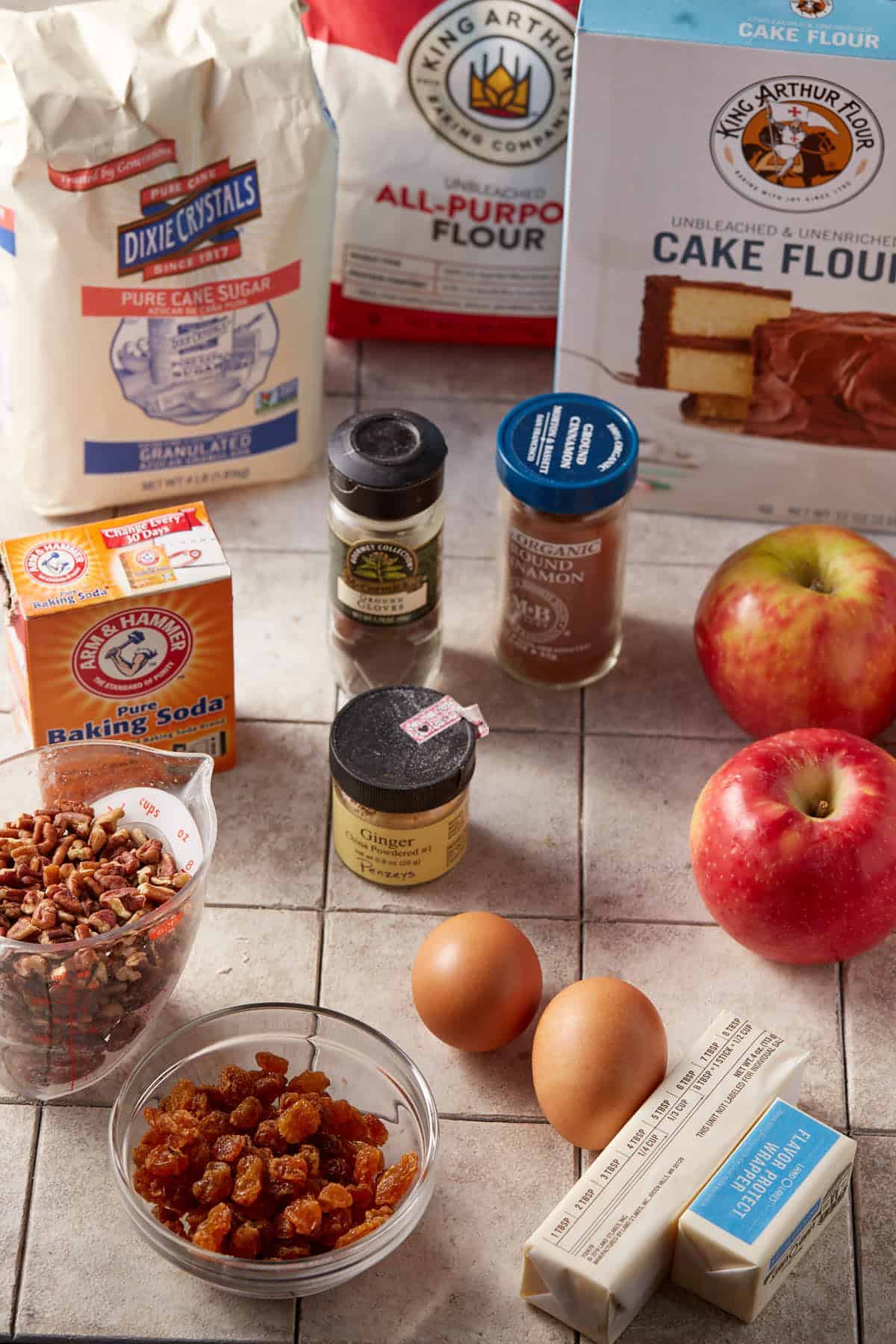 Baking ingredients for Apple Cinnamon Cake on a countertop: a bag of sugar, two types of flour, baking soda, spices, apples, pecans, golden raisins in a bowl, two eggs, and two sticks of butter.