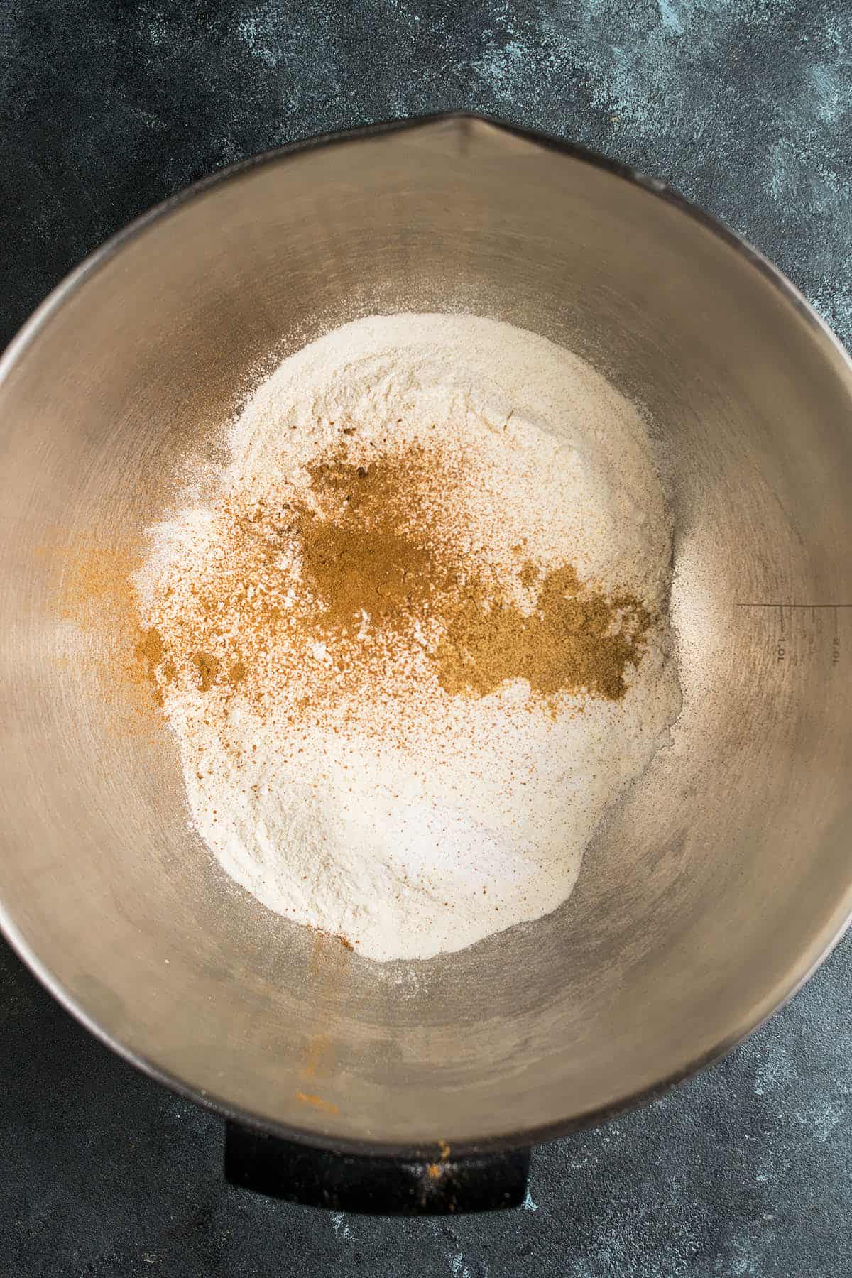 A metal mixing bowl containing flour and brown spices, possibly cinnamon or nutmeg, sits on a dark textured surface—perfect ingredients for baking a delicious Apple Cinnamon Cake.