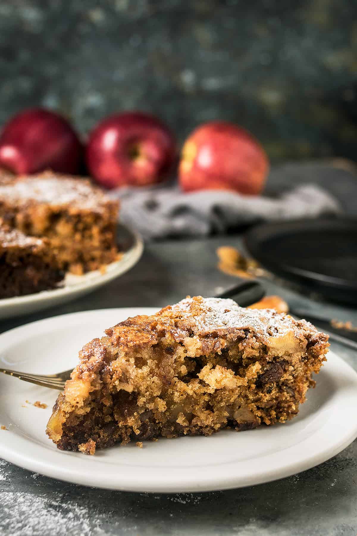 A close-up of a slice of moist Apple Cinnamon Cake dusted with powdered sugar on a white plate, with a fork beside it. Whole red apples and another cake slice are blurred in the background.