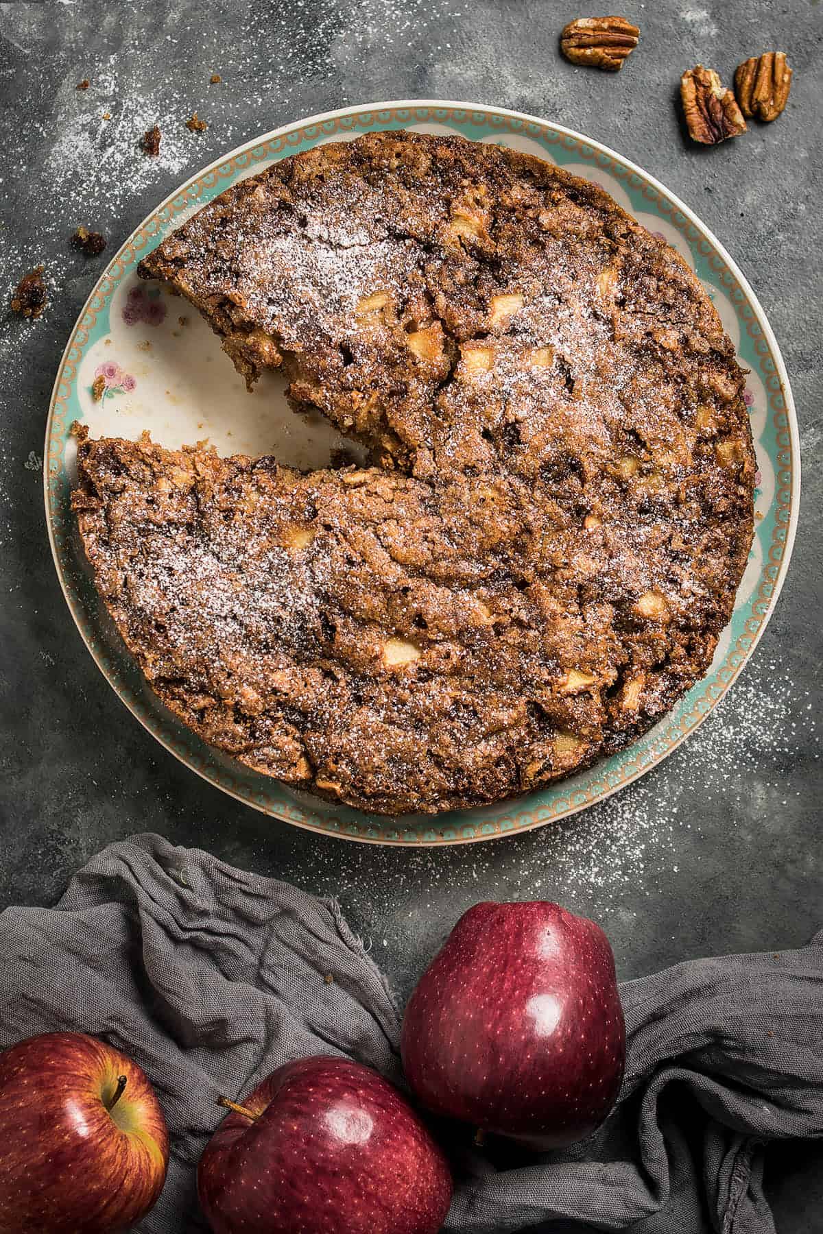 A rustic Apple Cinnamon Cake with a slice missing sits on a decorative plate, dusted with powdered sugar. Whole apples and pecans are nearby, with a gray cloth on a dark textured surface.