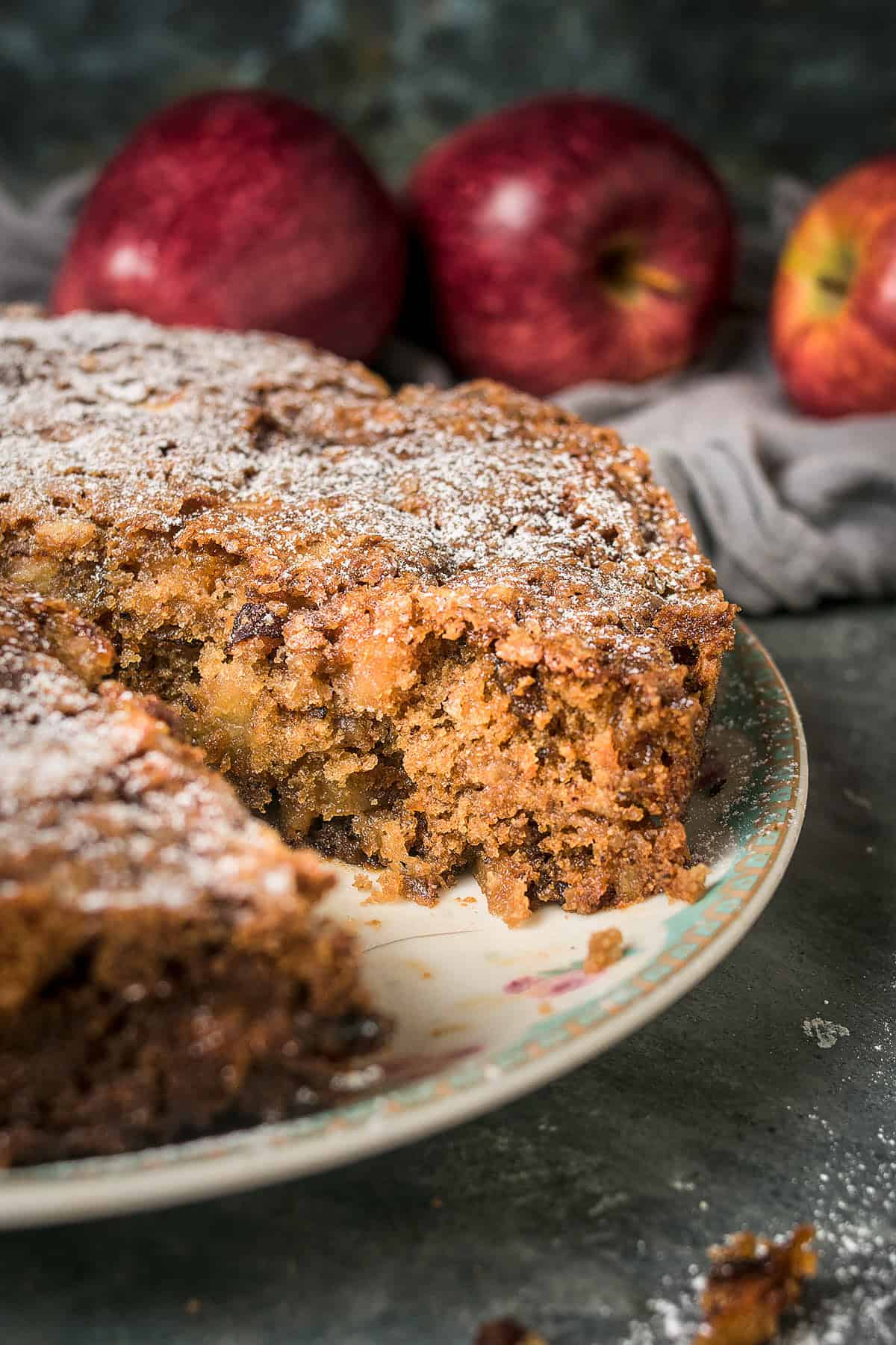 A close-up of a sliced Apple Cinnamon Cake on a decorative plate, topped with powdered sugar. Three whole red apples are blurred in the background on a dark surface.