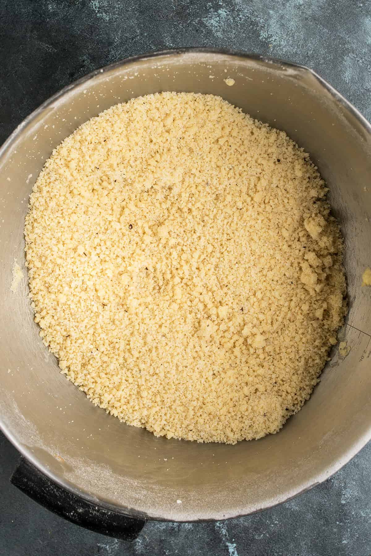 A metal mixing bowl filled with a coarse, crumbly mixture of flour and butter—perfect for starting an Apple Cinnamon Cake—rests on a dark countertop.