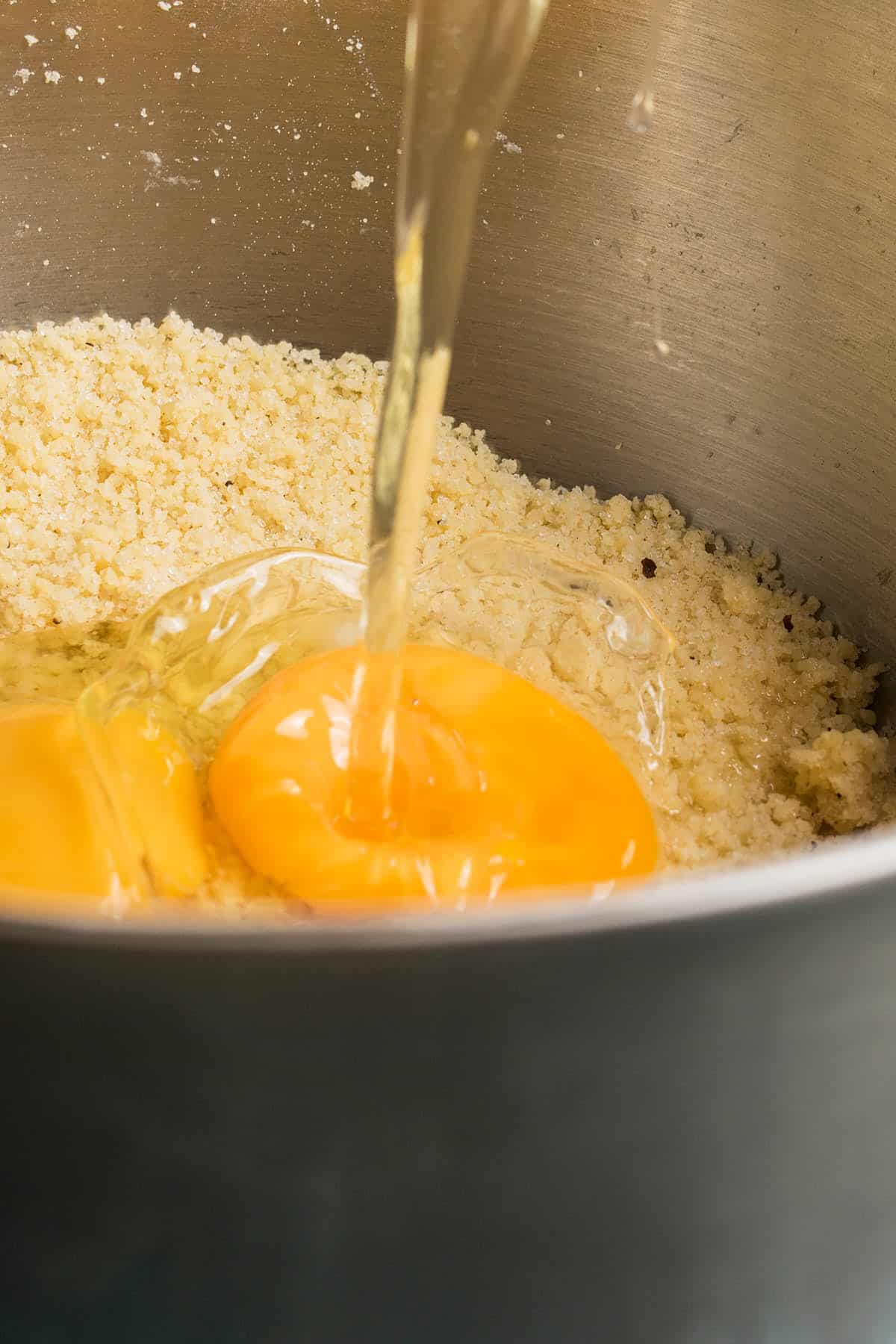 A close-up of raw eggs being poured into a metal mixing bowl filled with coarse, crumbly flour or dough mixture for an Apple Cinnamon Cake.