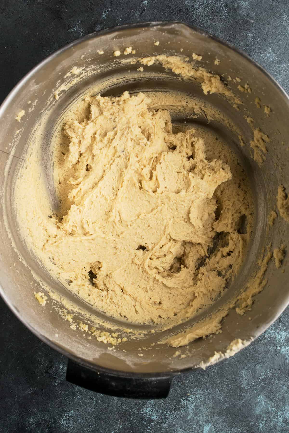 A metal mixing bowl filled with light brown Apple Cinnamon Cake dough, shown from above on a dark textured surface.