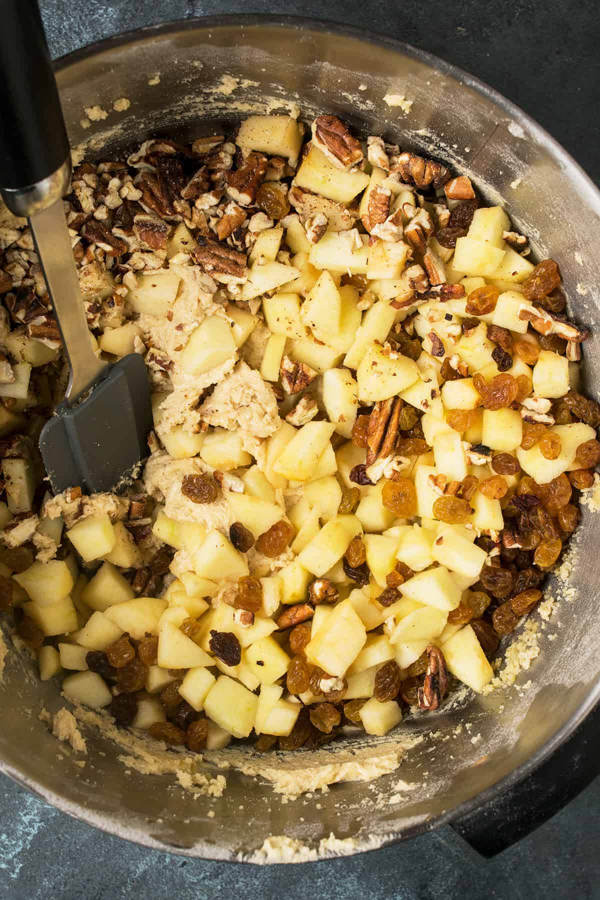 A mixing bowl filled with chopped apples, golden raisins, pecans, and dough being stirred with a black spatula for a delicious Apple Cinnamon Cake.