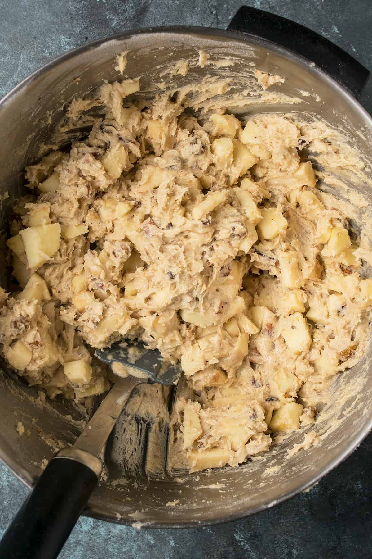 A mixing bowl filled with thick, lumpy Apple Cinnamon Cake dough containing visible chunks of apple is being mixed with a black spatula. The surface below is a dark, textured countertop.