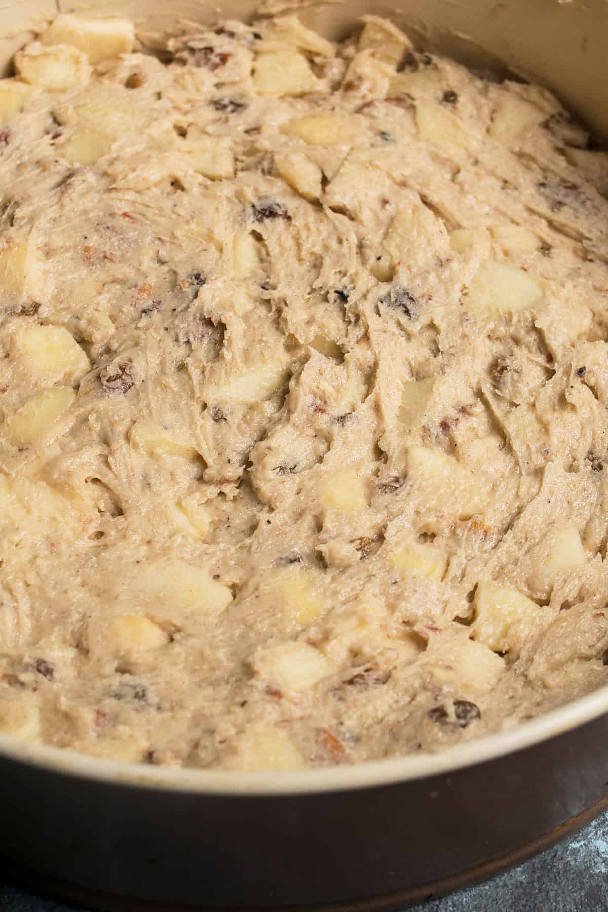 Close-up of an unbaked Apple Cinnamon Cake batter in a round baking pan, showing a thick mixture with visible chunks of apple and specks of nuts or raisins throughout.