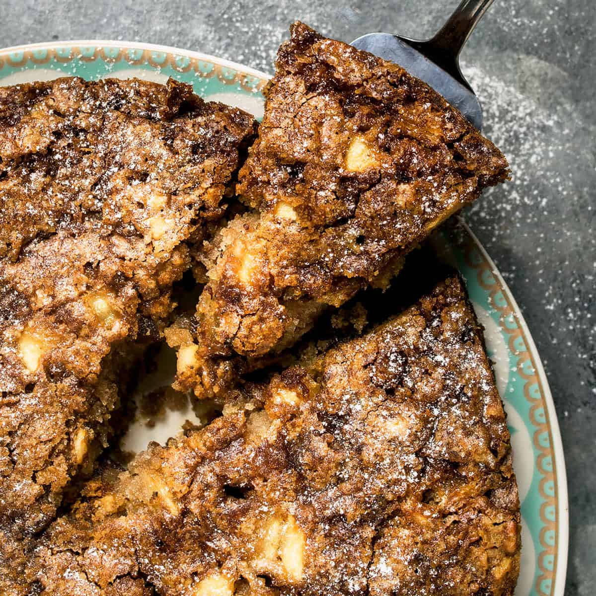 A close-up of a crumbly, golden-brown Apple Cinnamon Cake being served from a decorative plate, with a dusting of powdered sugar on top.